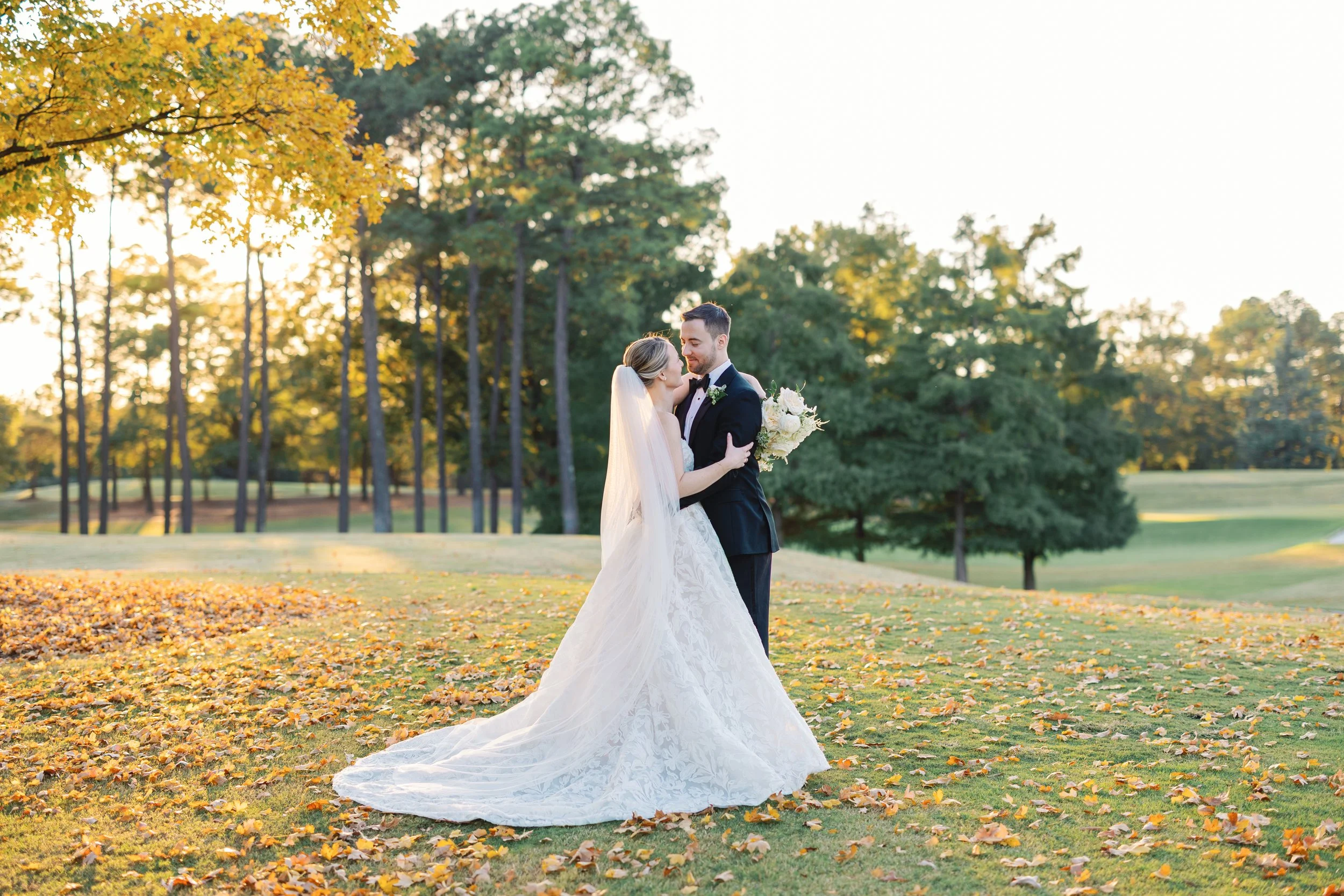 A bride and groom embrace on a grassy field with fallen leaves, tall trees, and a sunset in the background during a wedding photoshoot.