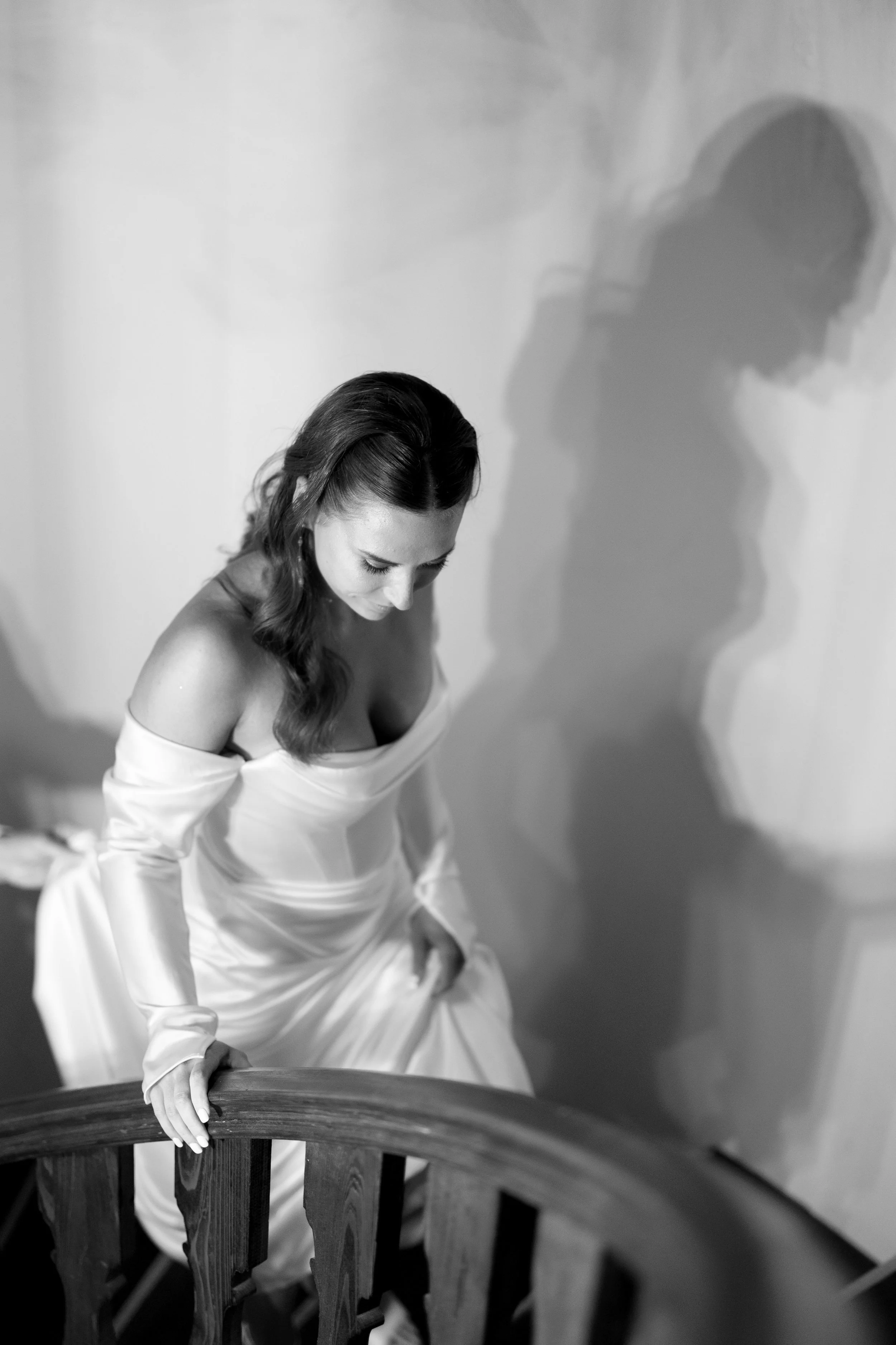 A woman in an off-the-shoulder white dress sitting on a staircase, looking down, with her shadow cast on the wall behind her.