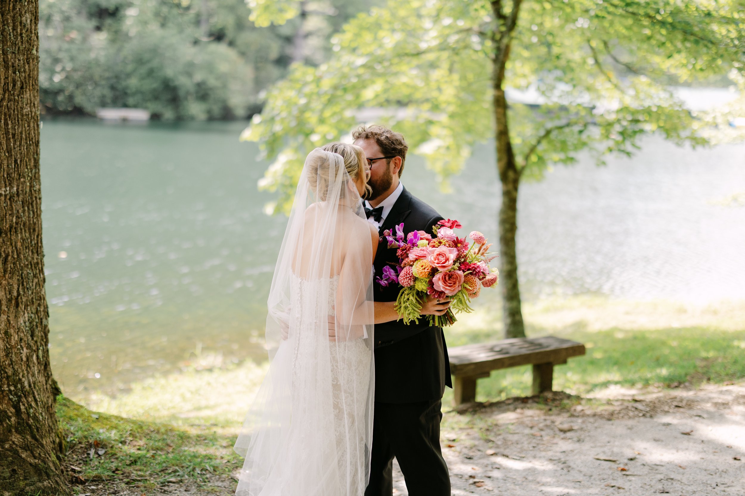 A bride and groom share a kiss outdoors near a lake, with trees and a park bench in the background. The bride wears a white wedding dress and veil, while the groom wears a black tuxedo with a bow tie. The groom holds a bouquet of pink and purple flow
