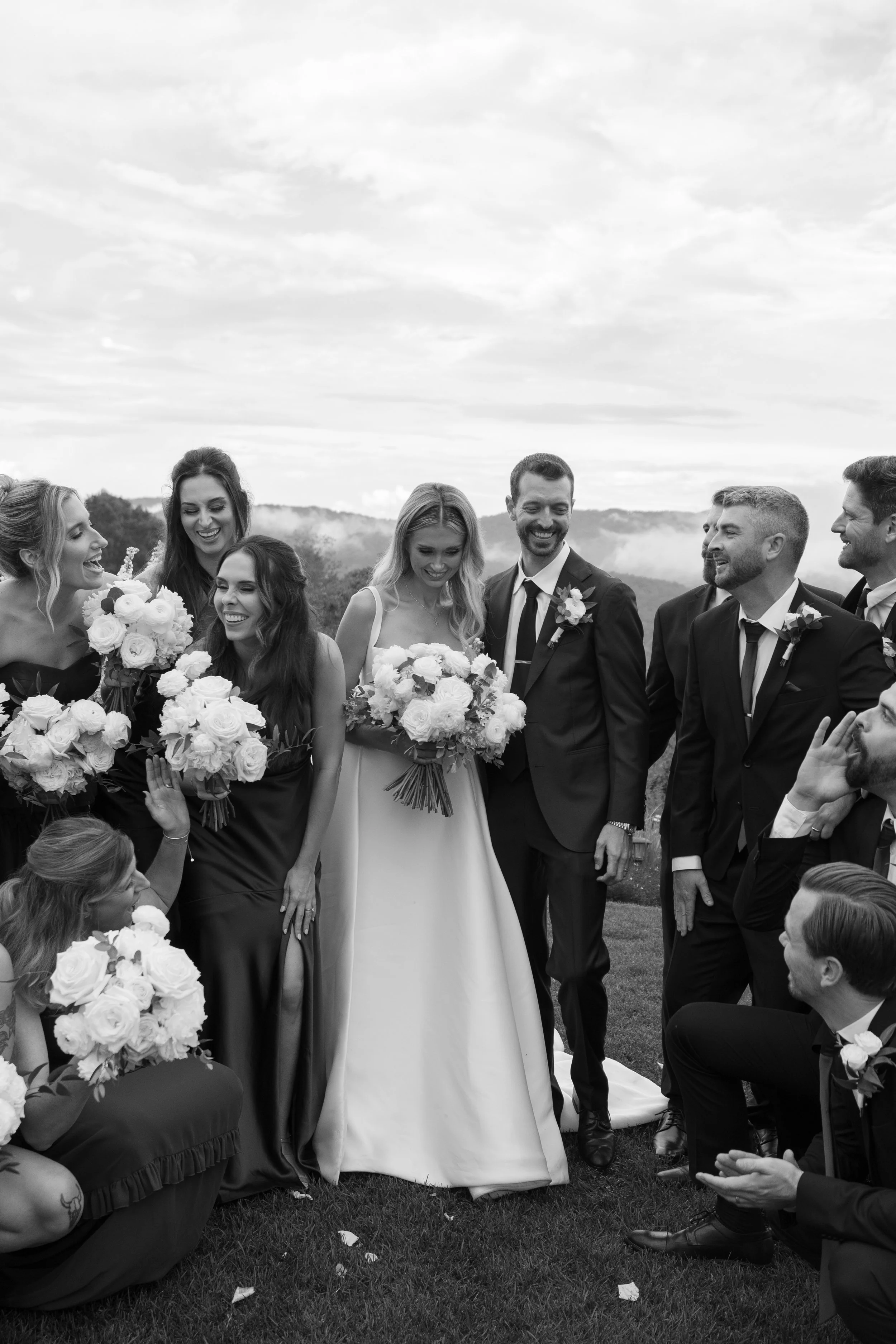 Black and white photo of a wedding celebration outdoors with the bride, groom, and wedding party smiling and holding bouquets.