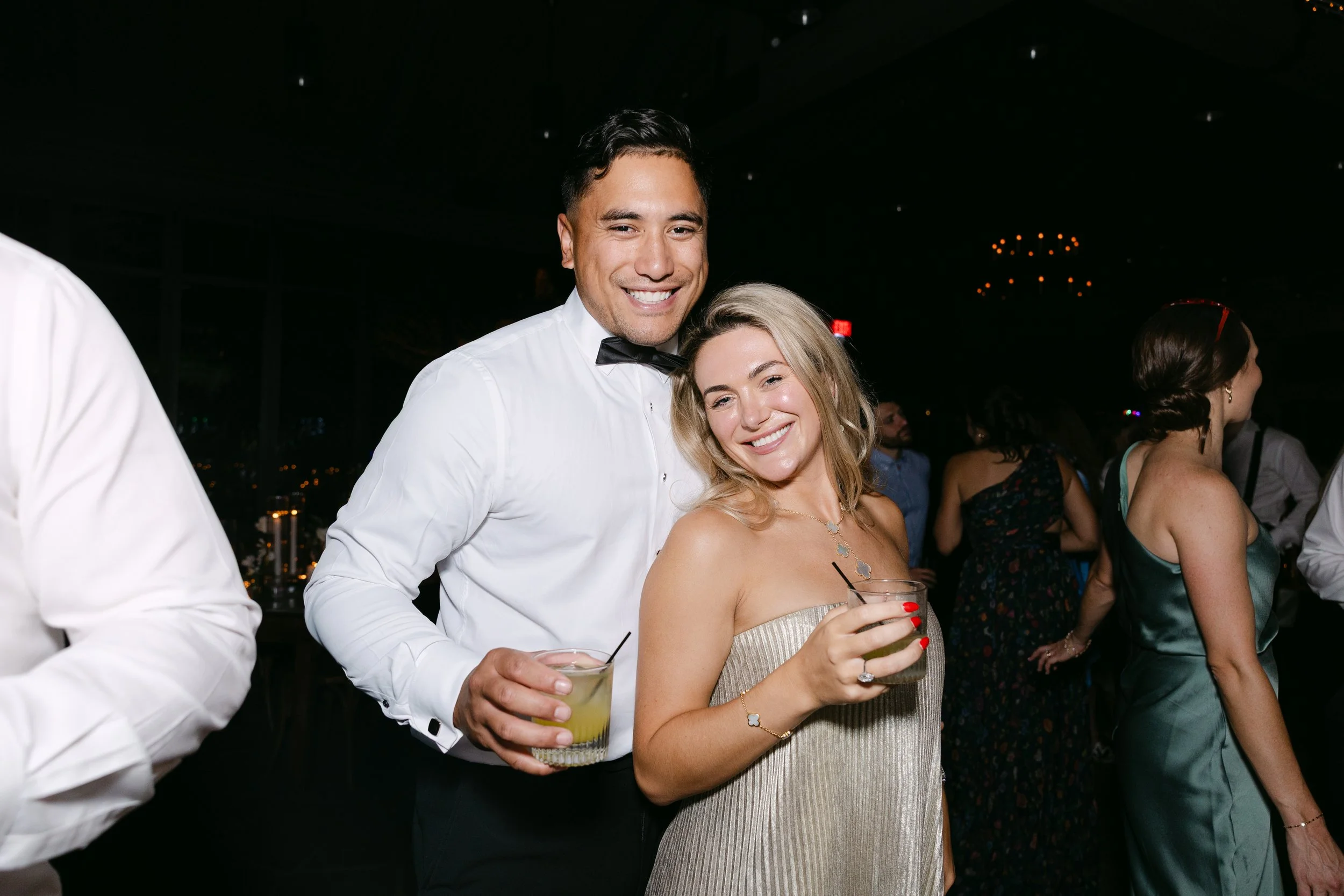 A man in a white dress shirt and black bow tie, and a woman in a strapless dress, smiling and holding drinks at a party.