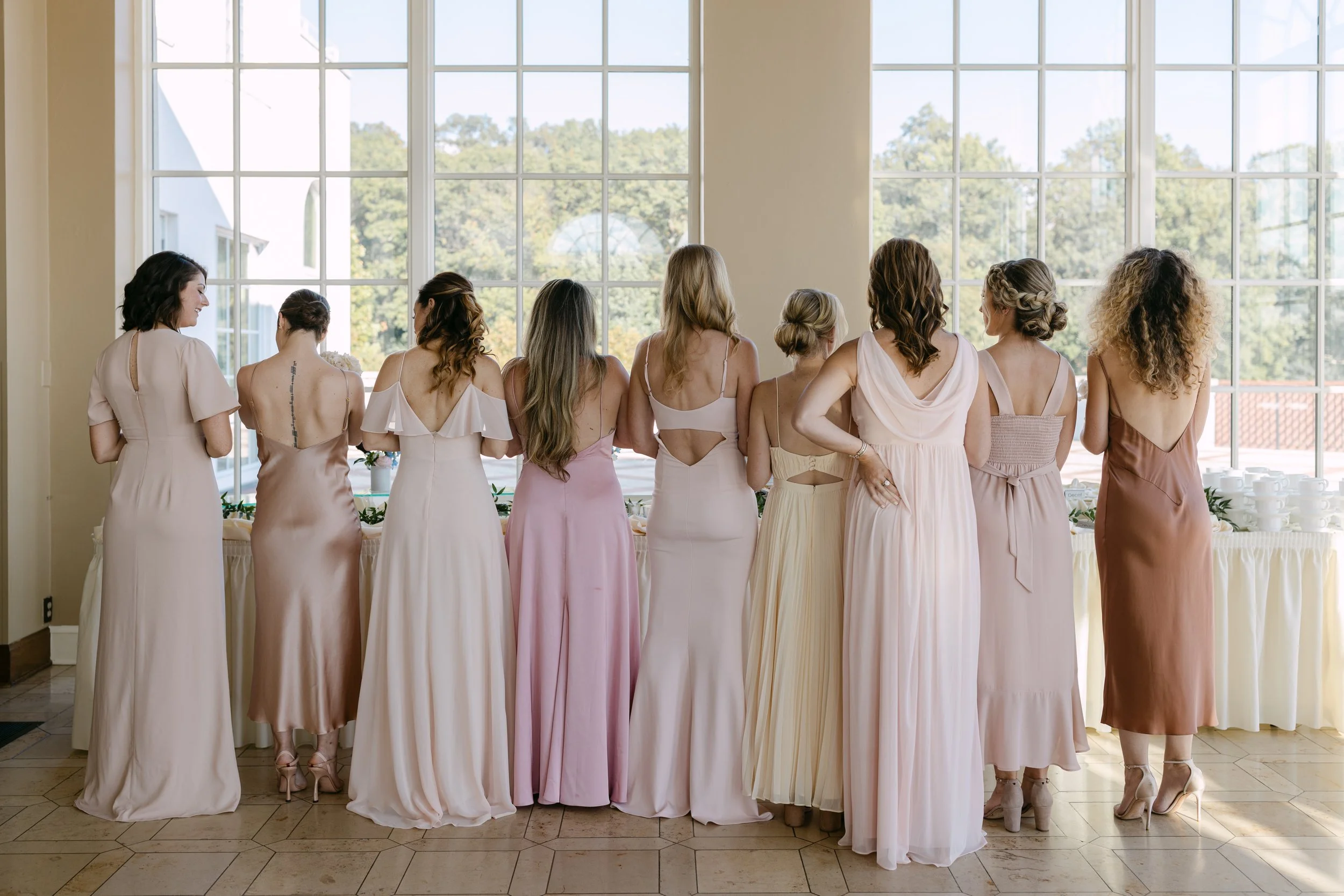 A group of women in pastel-colored dresses stand in a line facing large windows with a scenic outdoor view.