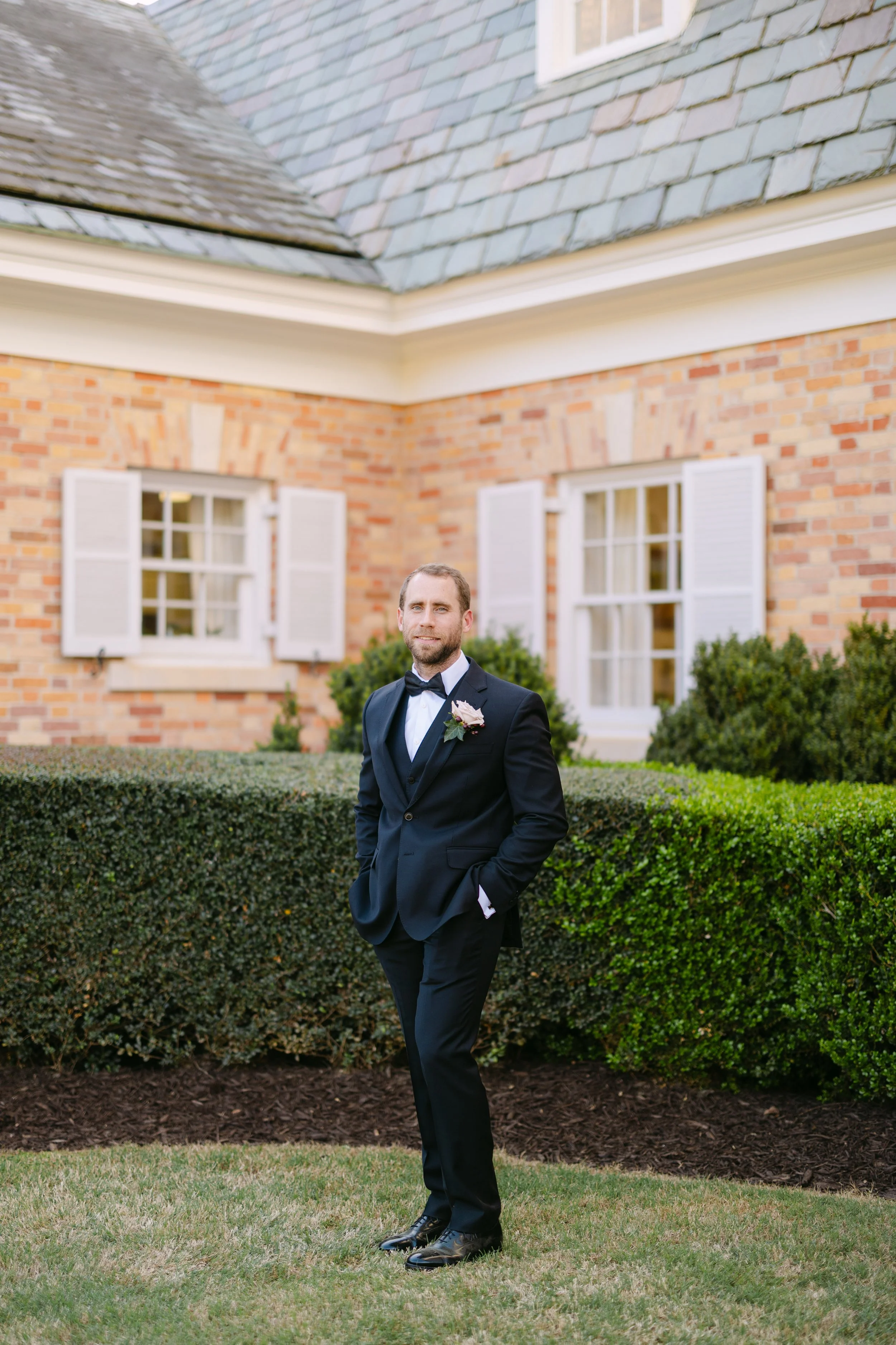 A man in a formal tuxedo with a boutonniere stands outdoors in front of a brick house with white window shutters and green bushes.