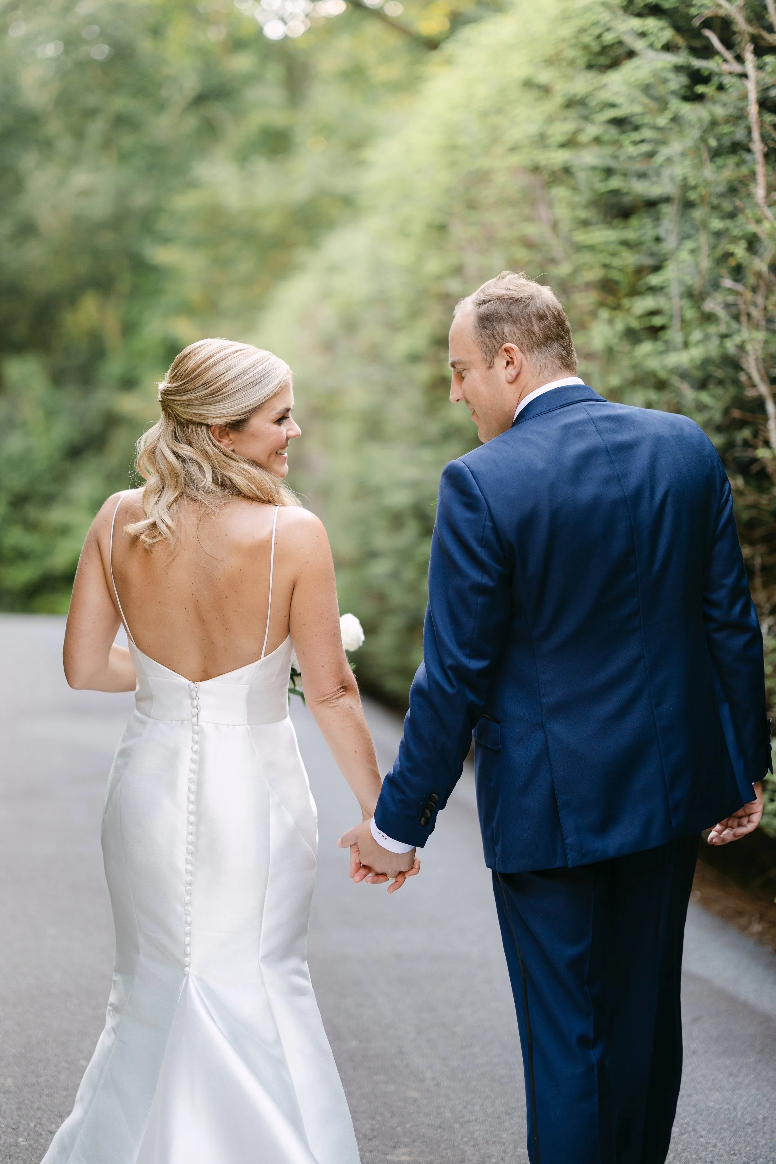 A bride and groom holding hands and looking at each other on a road surrounded by greenery.