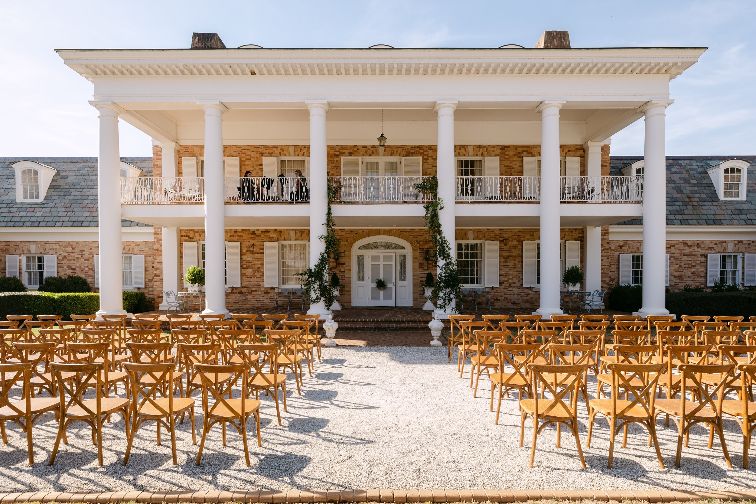 Outdoor wedding setup with wooden chairs arranged in front of a large two-story brick house with white columns and balcony, under clear sky.