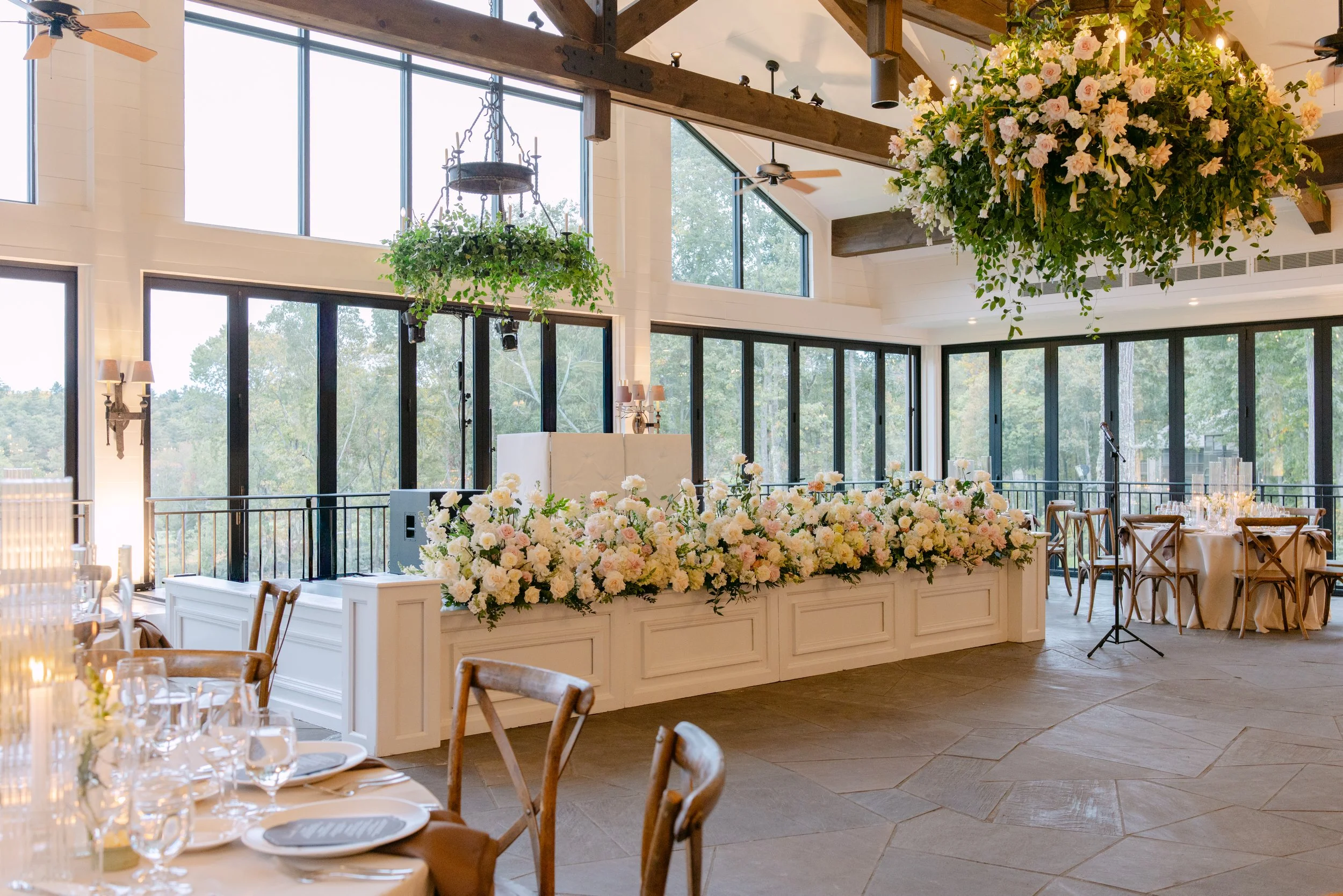 Elegant wedding reception venue with abundant white and blush floral arrangements, dining tables, exposed wood beams, and large windows overlooking trees