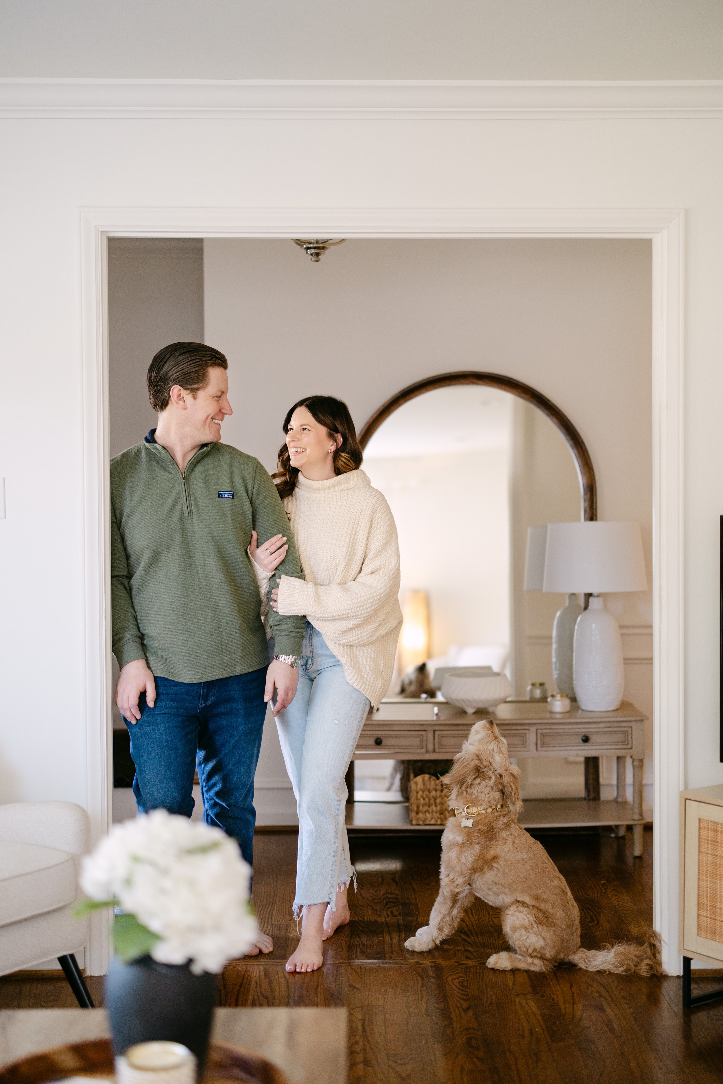 A couple walking together indoors, smiling and enjoying each other's company, with a golden retriever puppy sitting on the wooden floor and looking up at them.