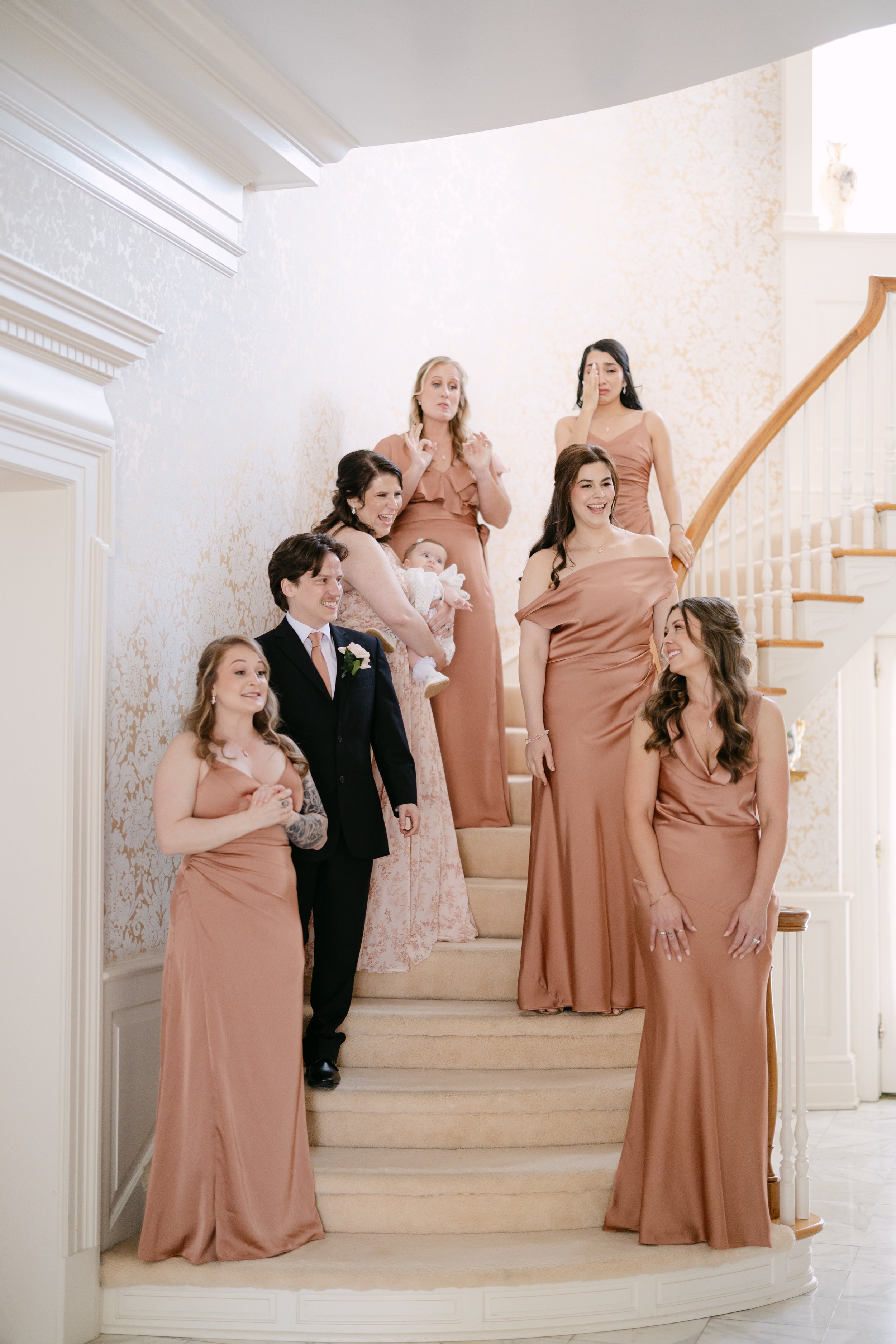 A group of women in matching peach-colored dresses and a man in a black suit with a pink tie posing on a curved staircase, likely at a wedding.