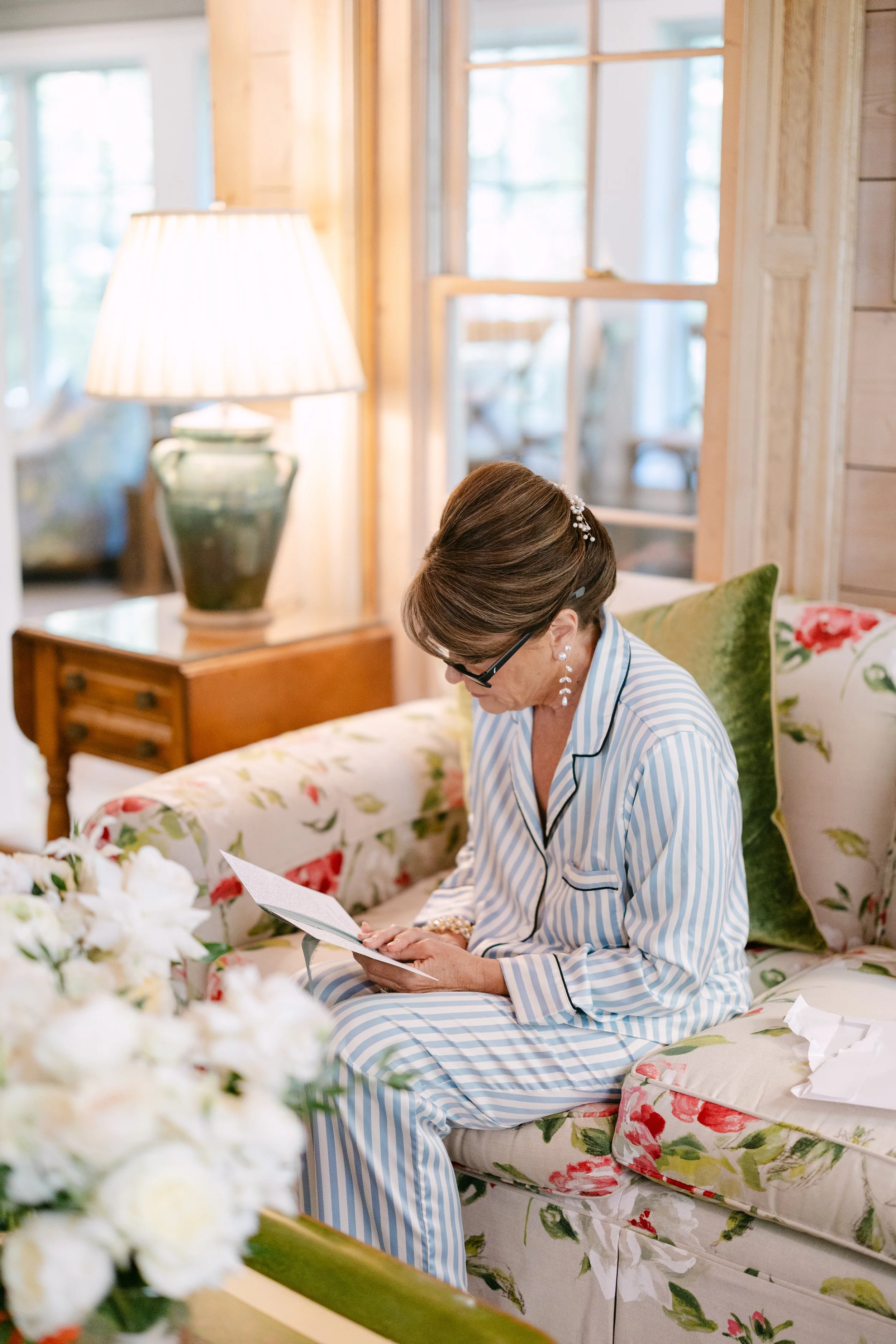 An elderly woman in blue and white striped pajamas sitting on a floral sofa, reading a card in a cozy, well-lit room with wooden walls and large windows.