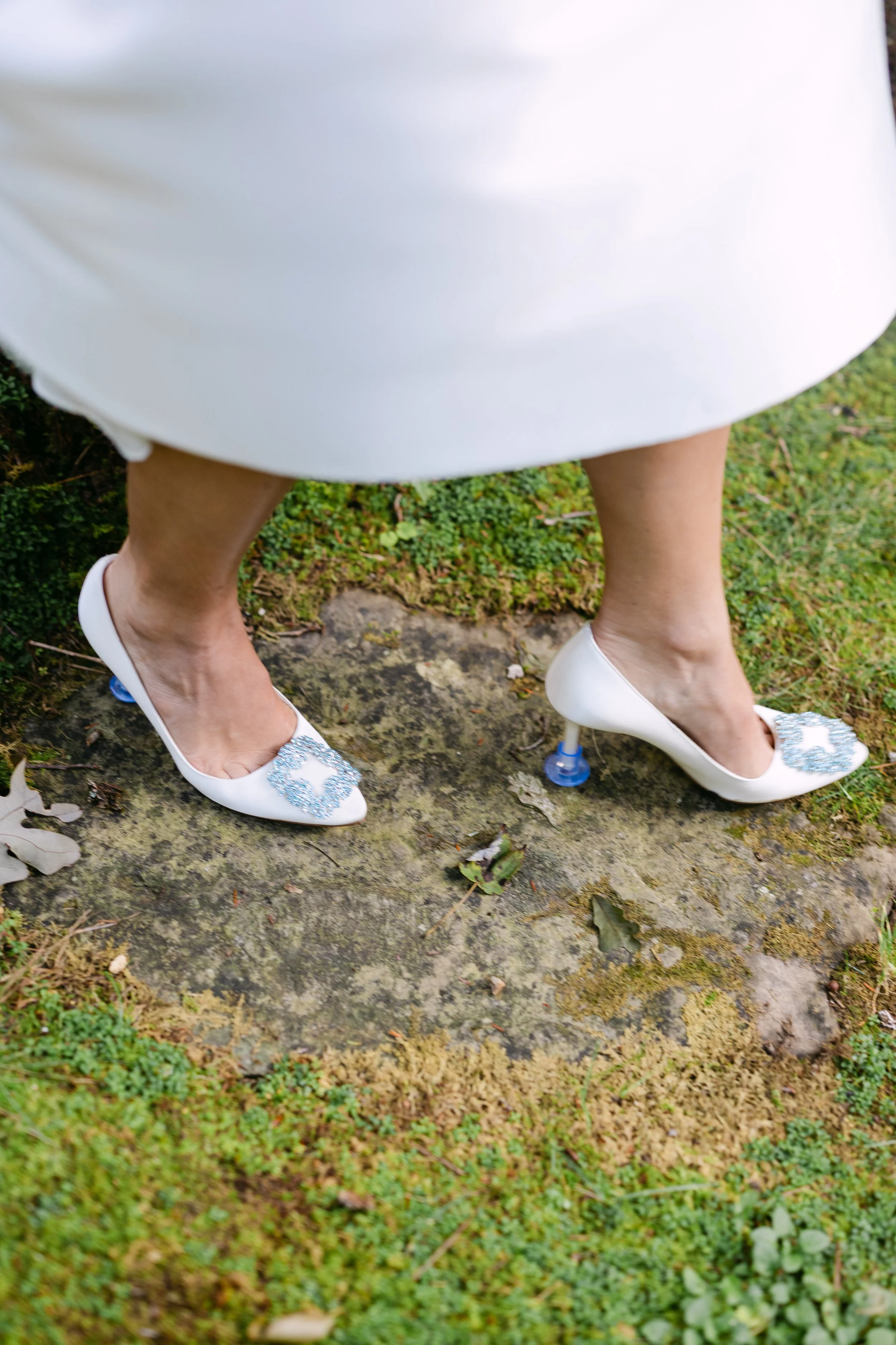 Close-up of a woman wearing white high-heeled shoes with blue circular embellishments and a white dress, standing outdoors on mossy ground.
