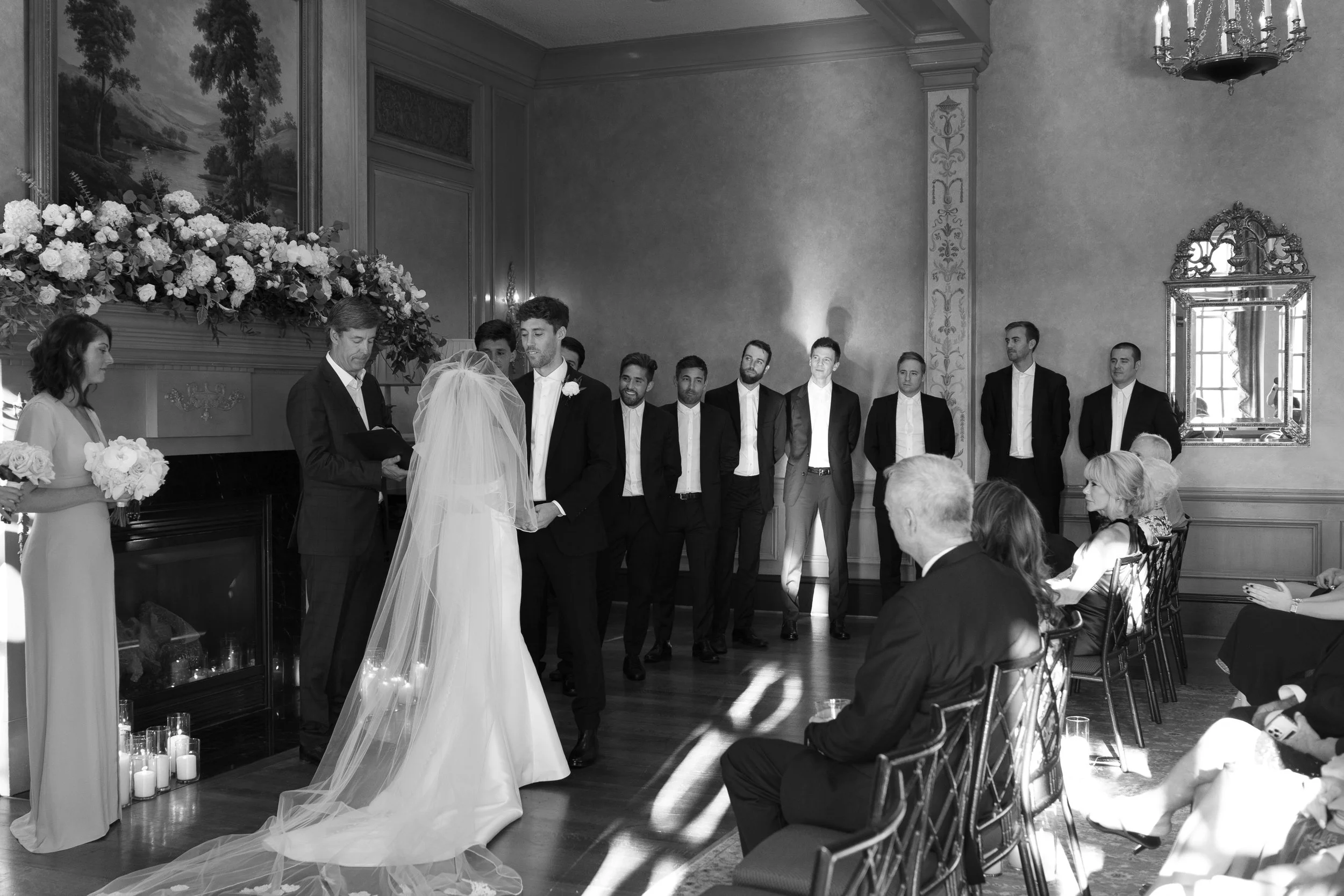 A black-and-white photo of a wedding ceremony with a bride and groom exchanging vows in front of an officiant, with bridesmaids and groomsmen standing behind them, in an elegant room with floral decorations and guests seated.