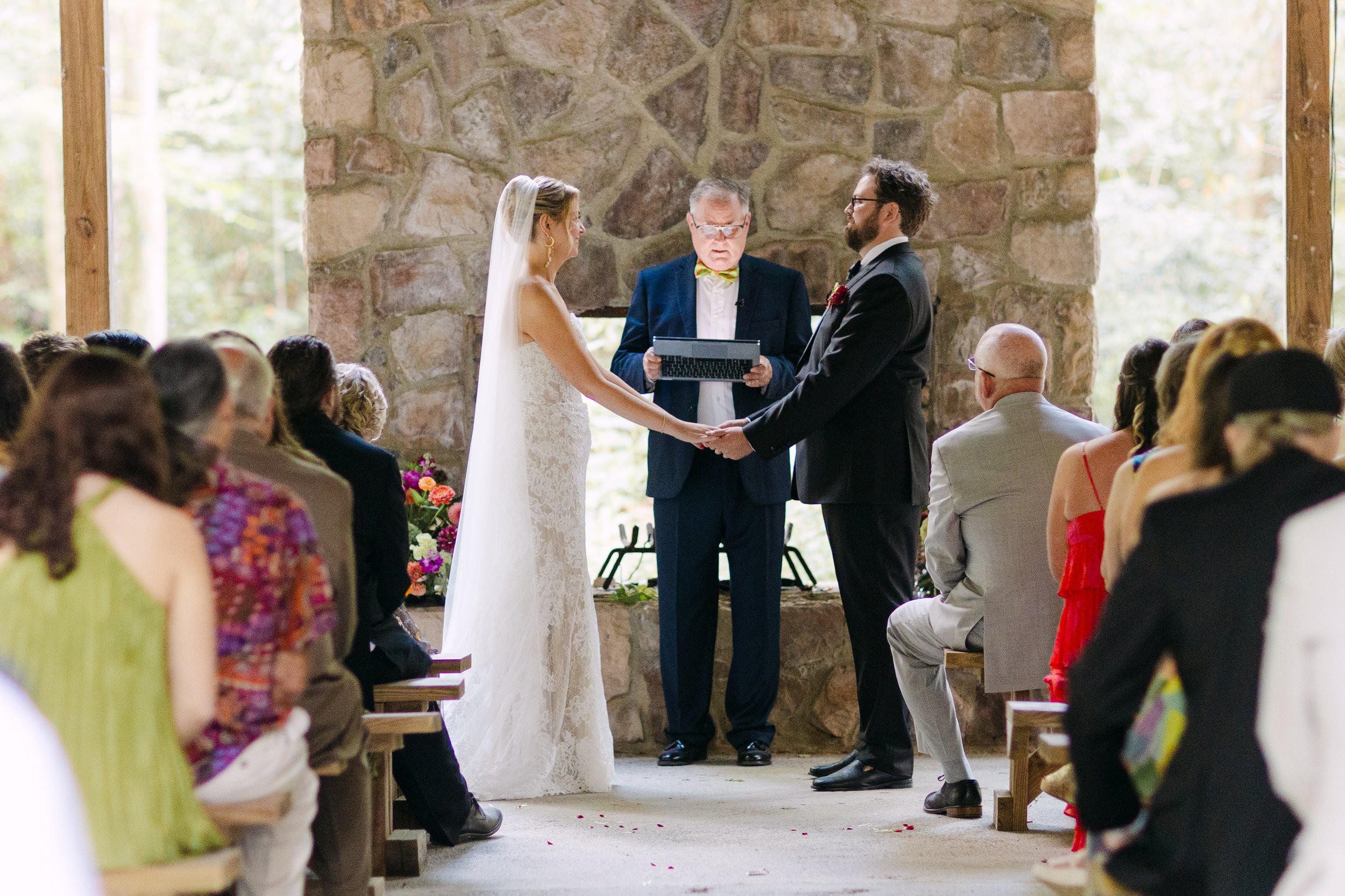 A wedding ceremony with a bride and groom holding hands, standing before an officiant, inside a rustic stone fireplace setting, surrounded by seated guests.