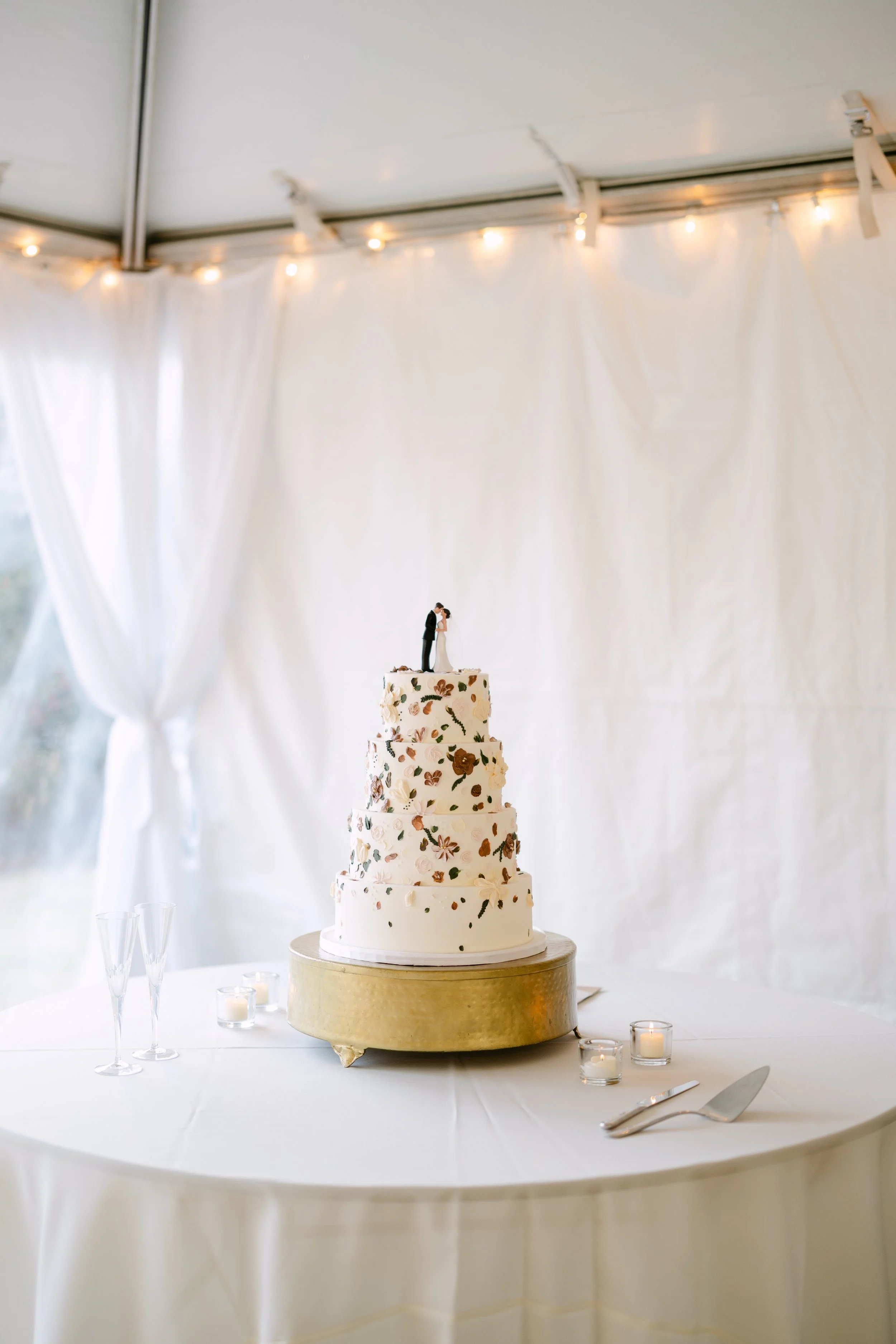 A multi-tiered wedding cake with floral decorations and a bride and groom topper, placed on a round table with candles and champagne glasses in a decorated venue.