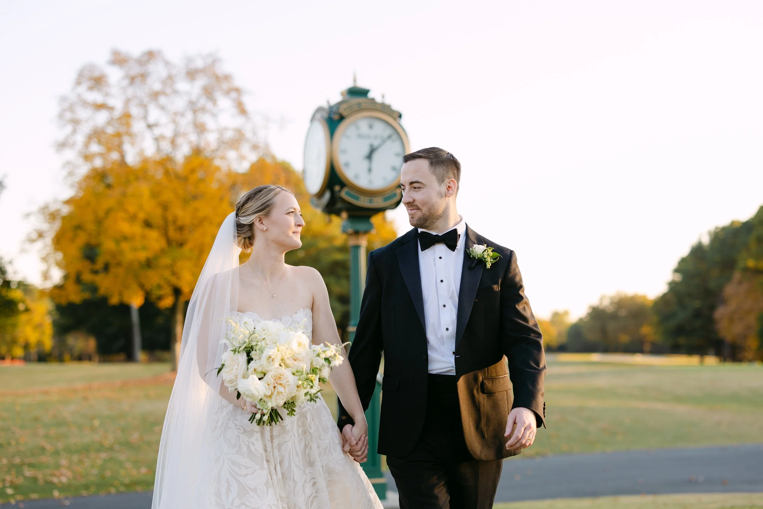 A bride and groom holding hands, walking outdoors during their wedding, with a clock tower and autumn trees in the background.
