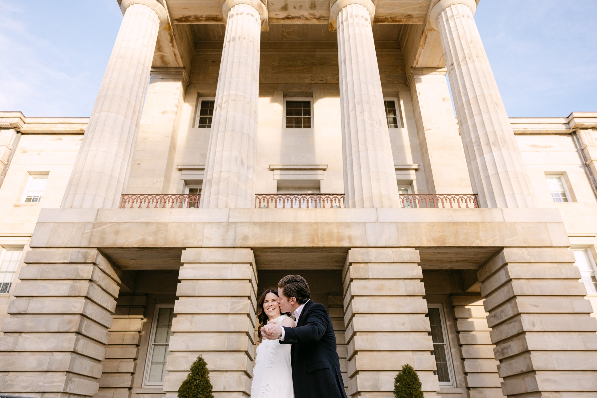 A newlywed couple dancing in front of a grand stone building with tall pillars and columns, under a clear sky.