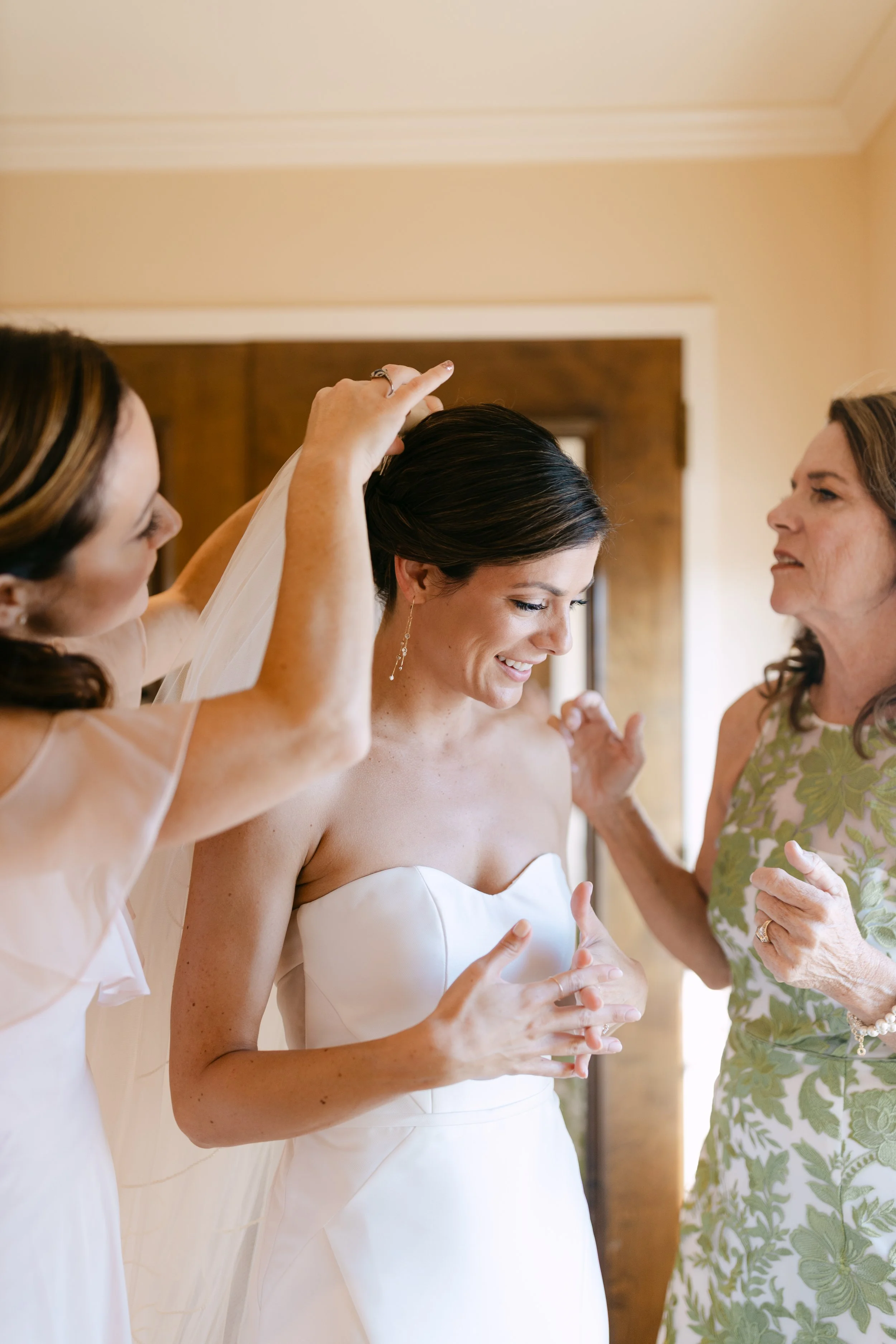 A bride smiling while getting ready with two women helping her adjust her veil in a room.