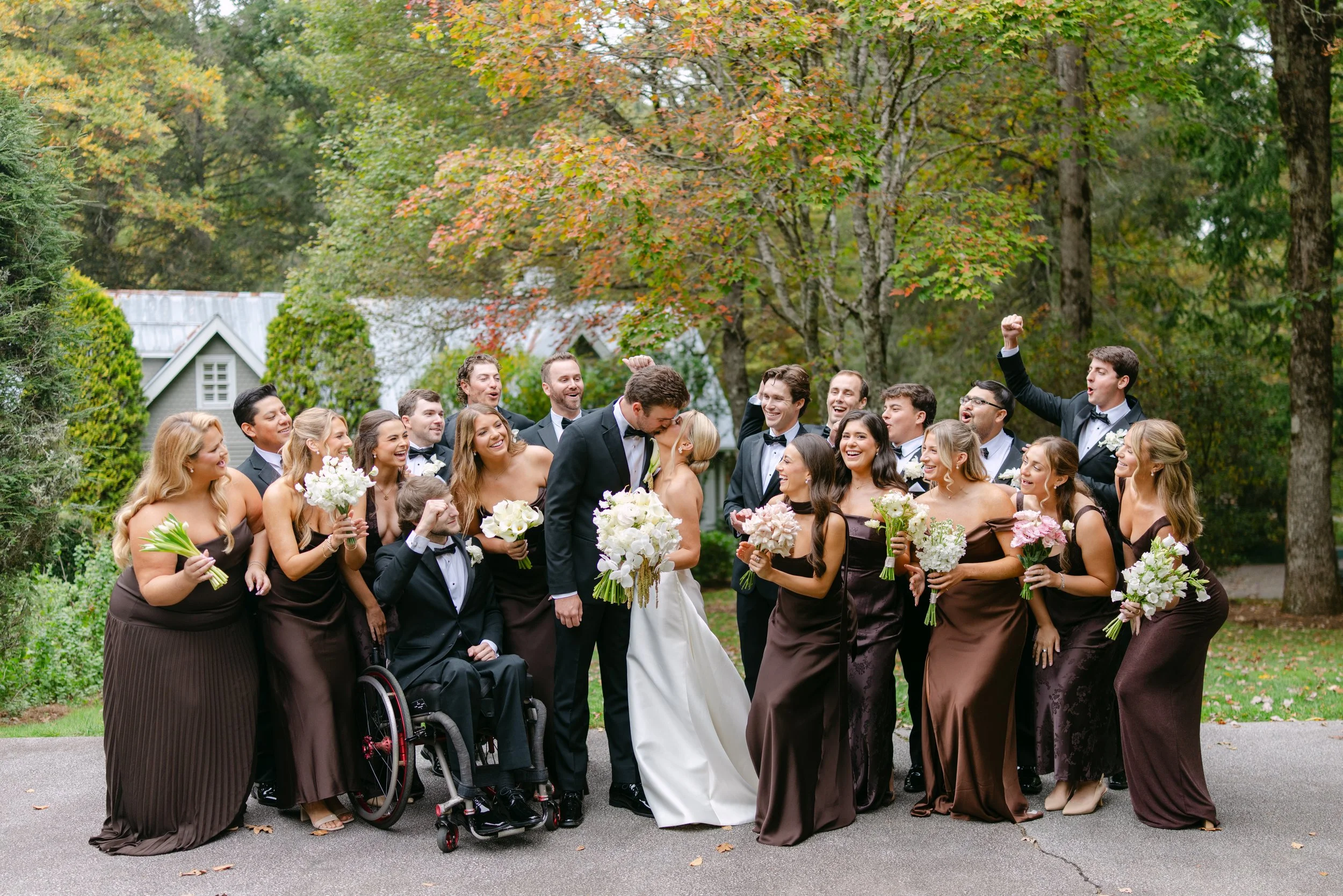 Joyful wedding party posing outdoors in autumn, with the bride, groom, bridesmaids holding bouquets, groomsmen, and a guest in a wheelchair.