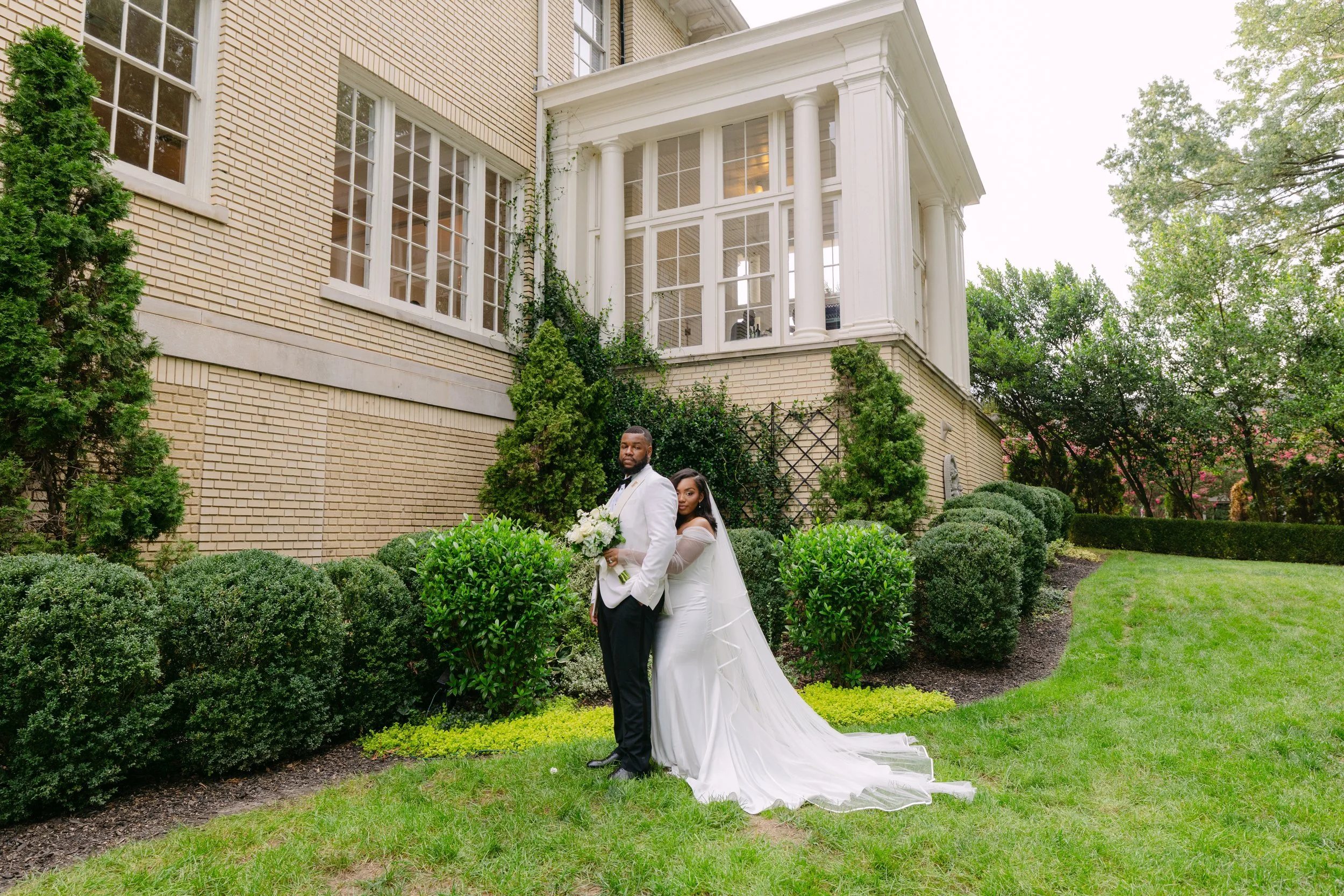 A newly married couple in wedding attire standing outdoors on a neatly landscaped garden lawn with green bushes, trees, and a large brick house with white trim and windows in the background.