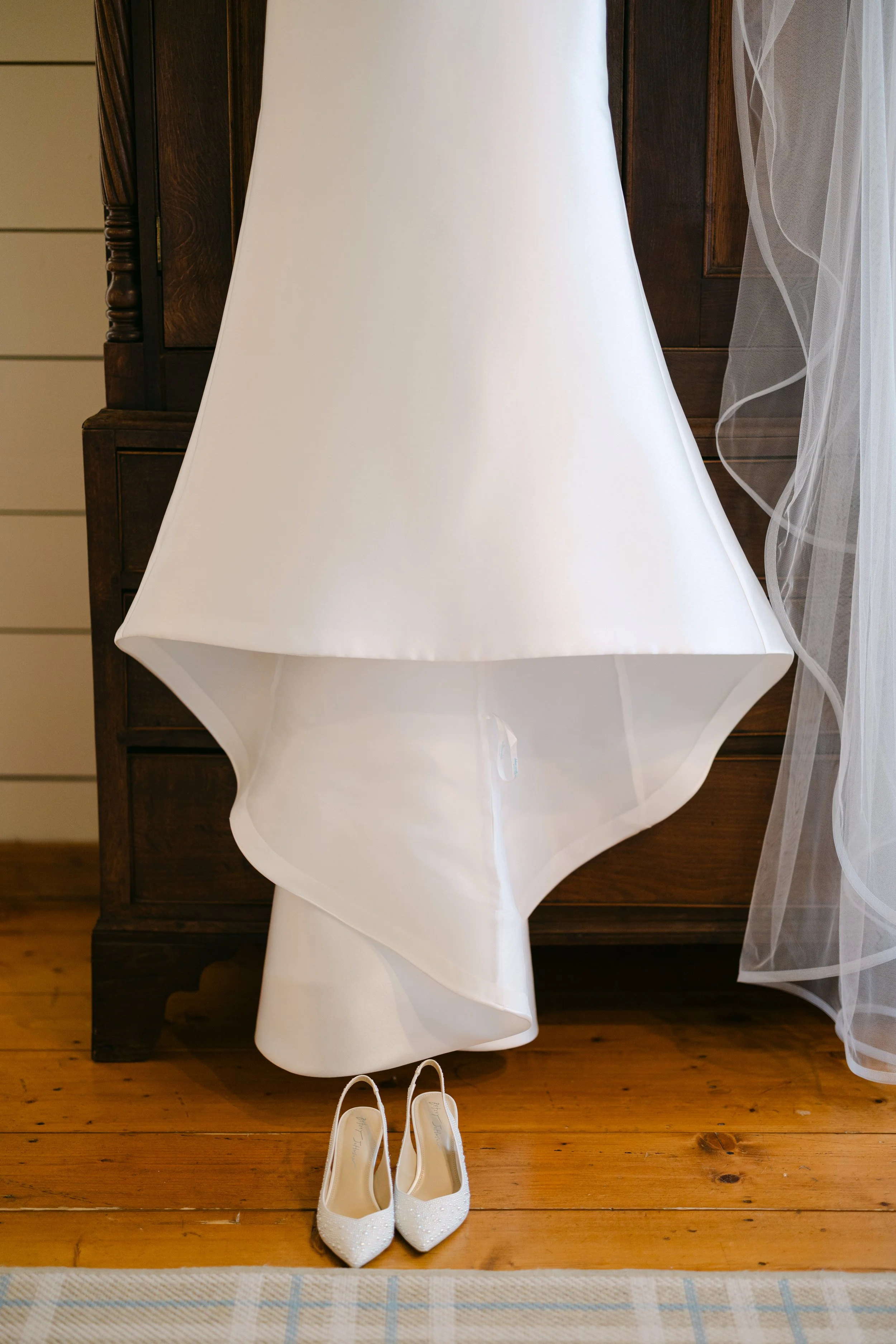 A white wedding dress hangs in front of a wooden dresser with a pair of white, glittery high-heeled shoes placed beneath it on a wooden floor.
