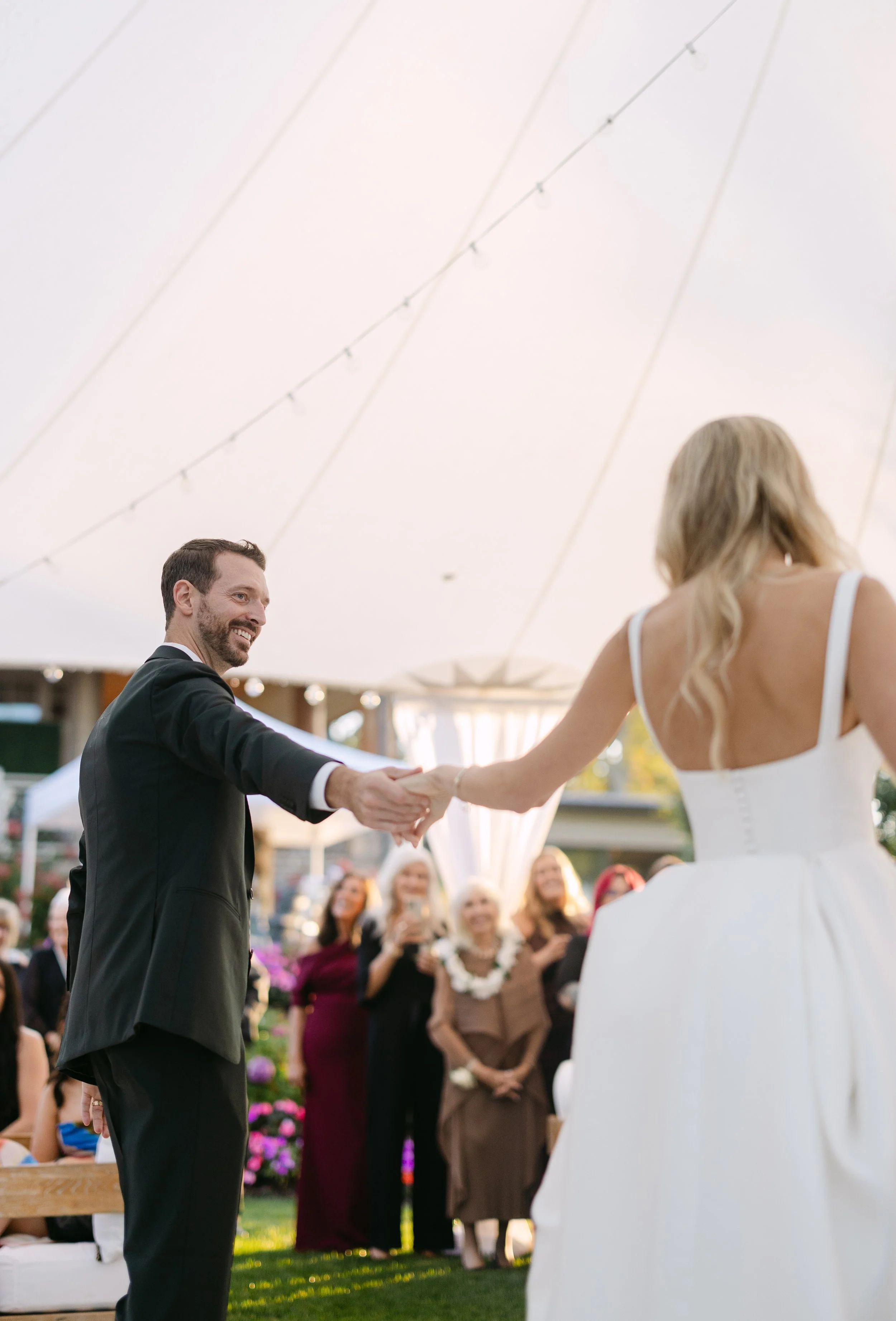 A couple is holding hands and smiling during a wedding ceremony under a large white tent, with guests watching in the background.