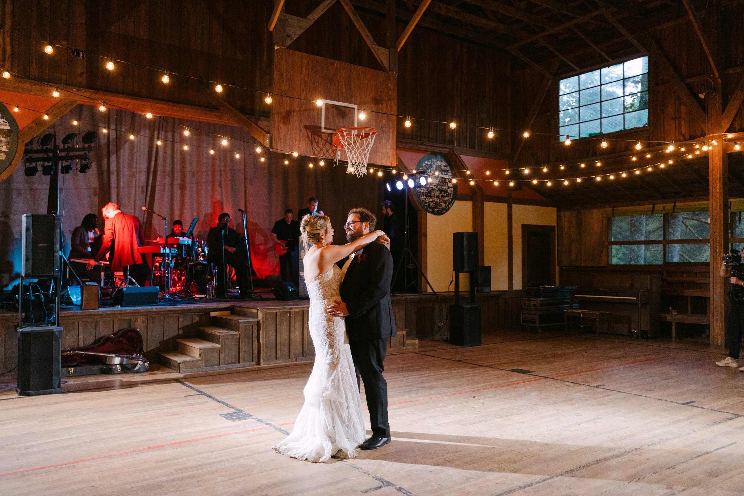 A bride and groom are dancing together in a rustic wooden barn with string lights overhead, with a live band playing on a stage in the background.