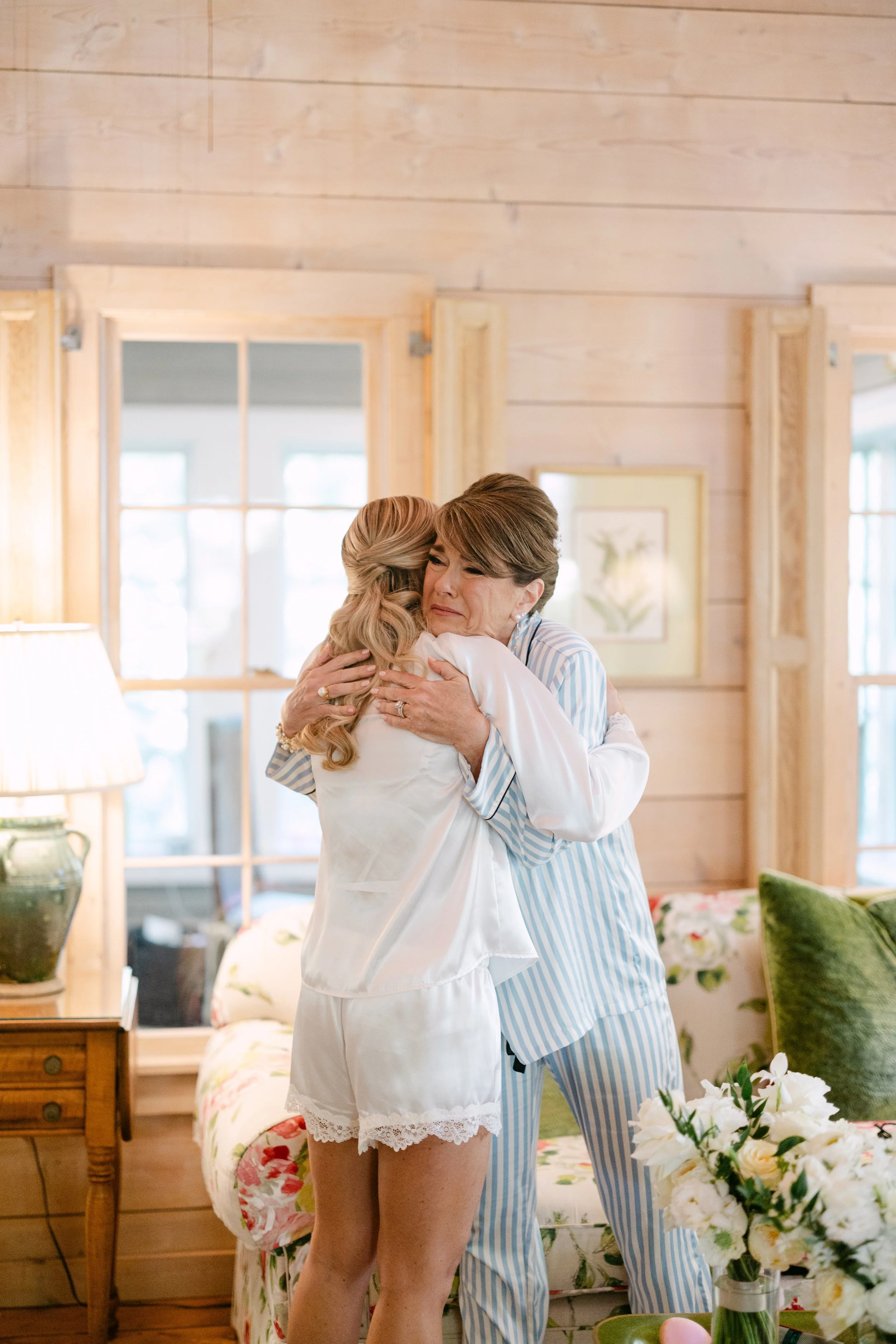 Two women hugging in a cozy, wooden-paneled room with floral decor and large windows in the background.