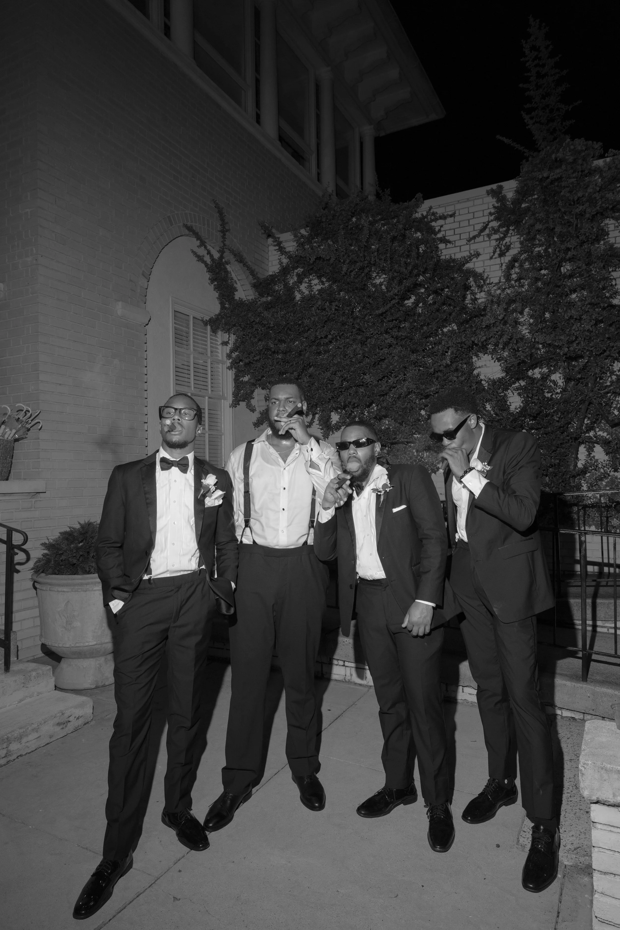 Five men dressed in formal suits with bow ties and sunglasses, standing outdoors at night in front of a building with brick and stone architecture, some smoking cigars.