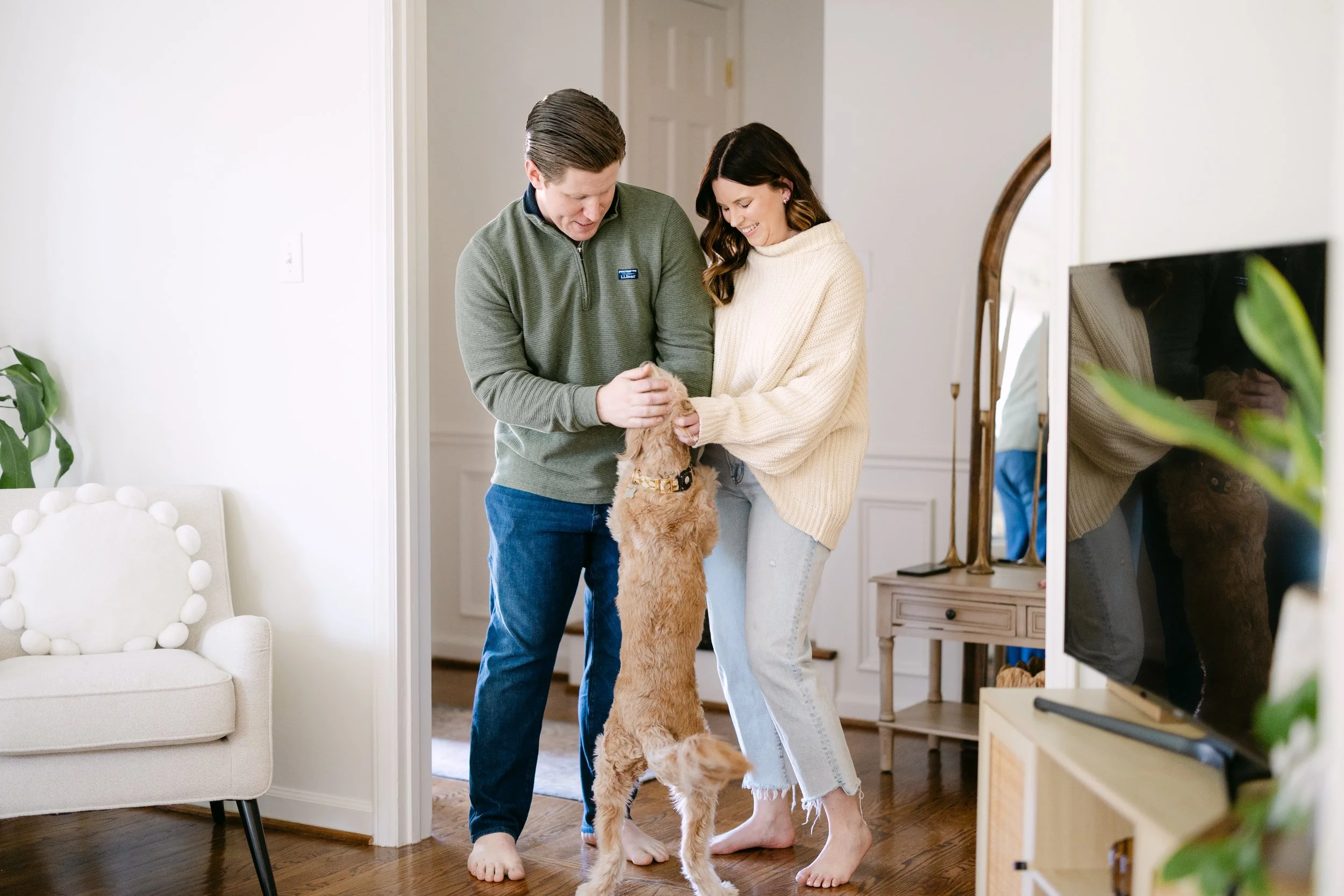 Two people, a man and a woman, playing with a golden retriever puppy indoors in a living room. The man is holding the puppy's paws, and the woman is smiling and holding the puppy's body. The puppy is standing on its hind legs.