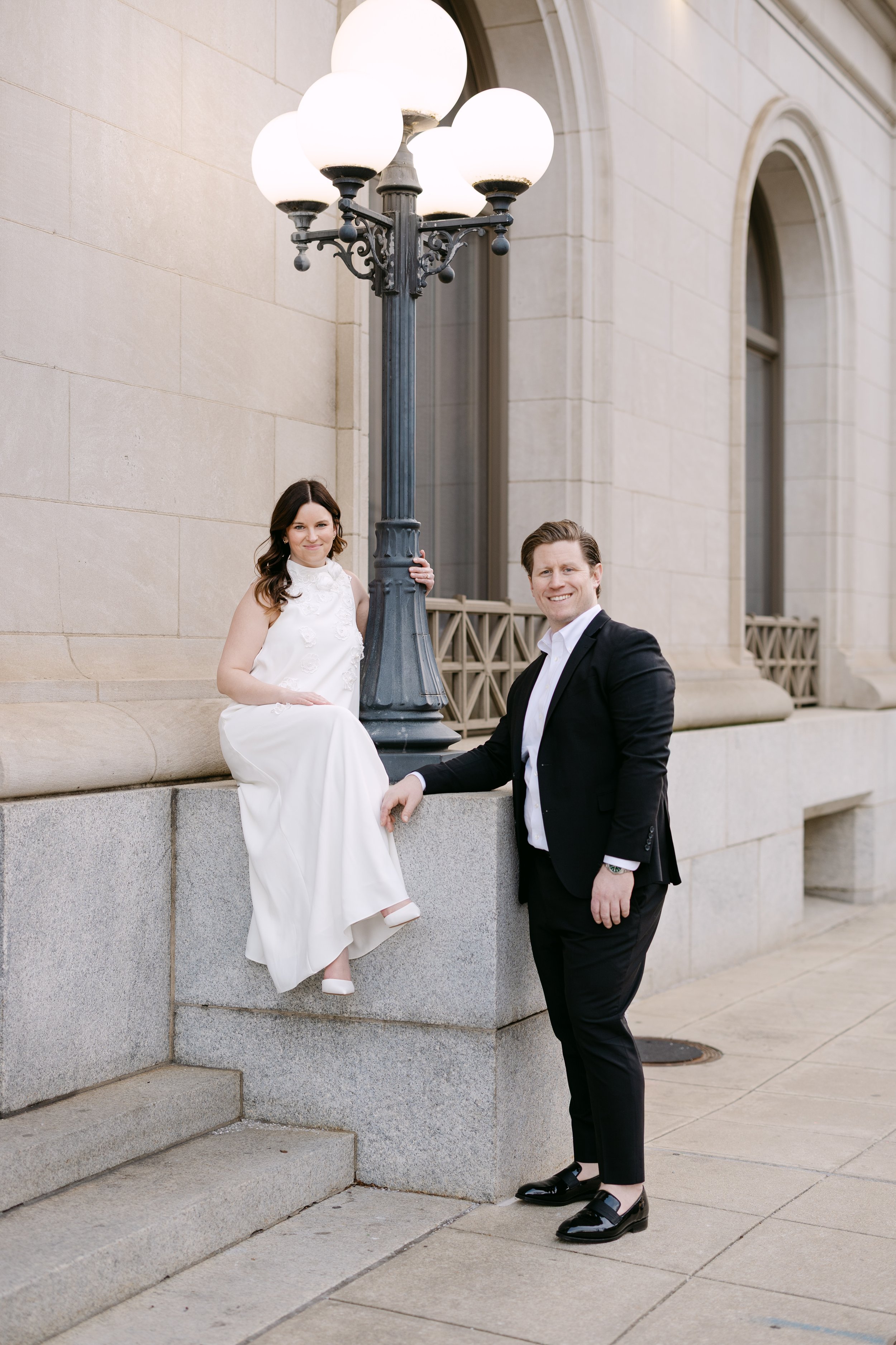 A woman in a white dress sitting on a stone ledge and a man in a black suit standing next to her, outdoors near a building with large windows, a streetlamp, and steps.