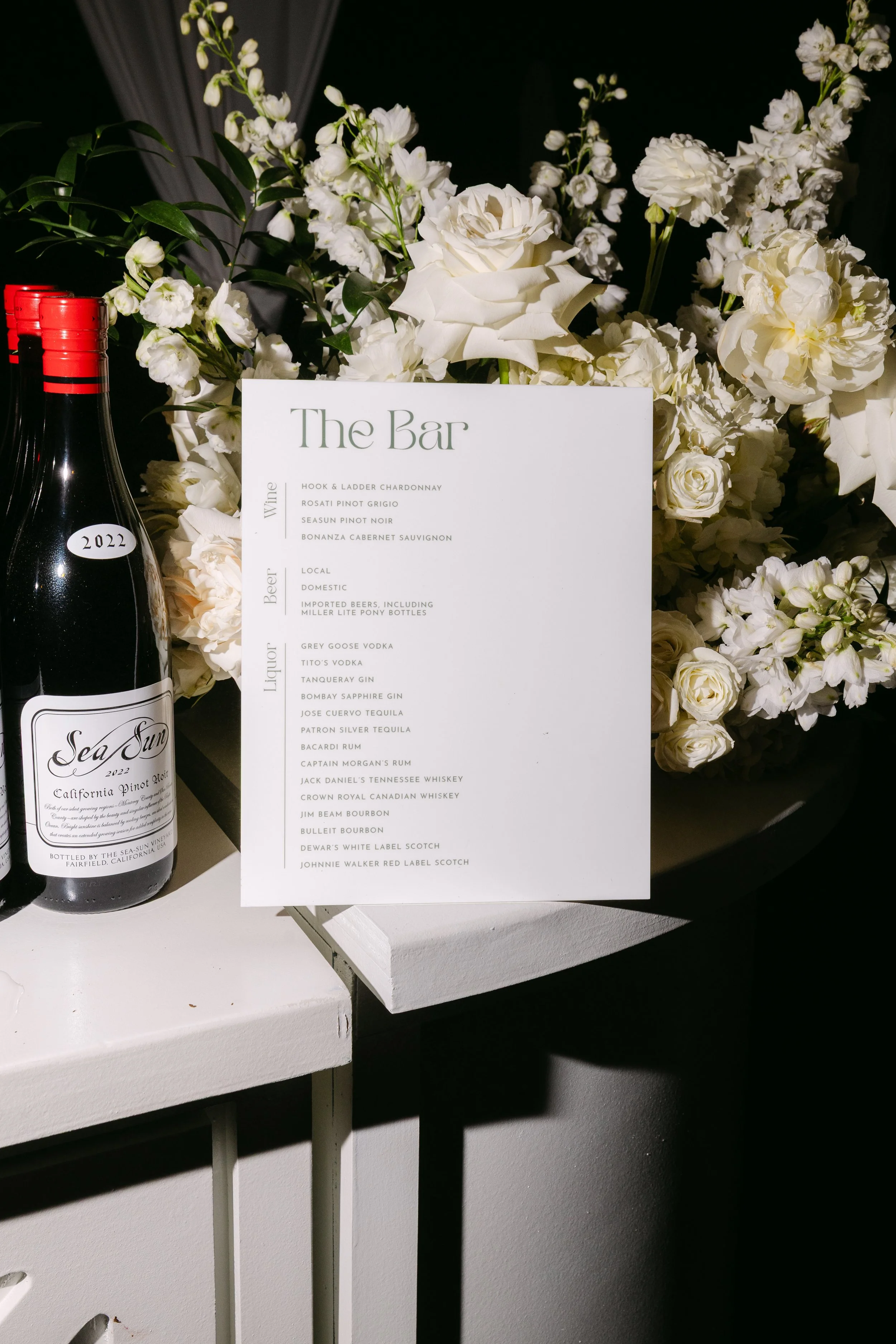 A white printed menu titled 'The Bar' surrounded by white flowers and a bottle of Sea Sun California Pinot Noir.