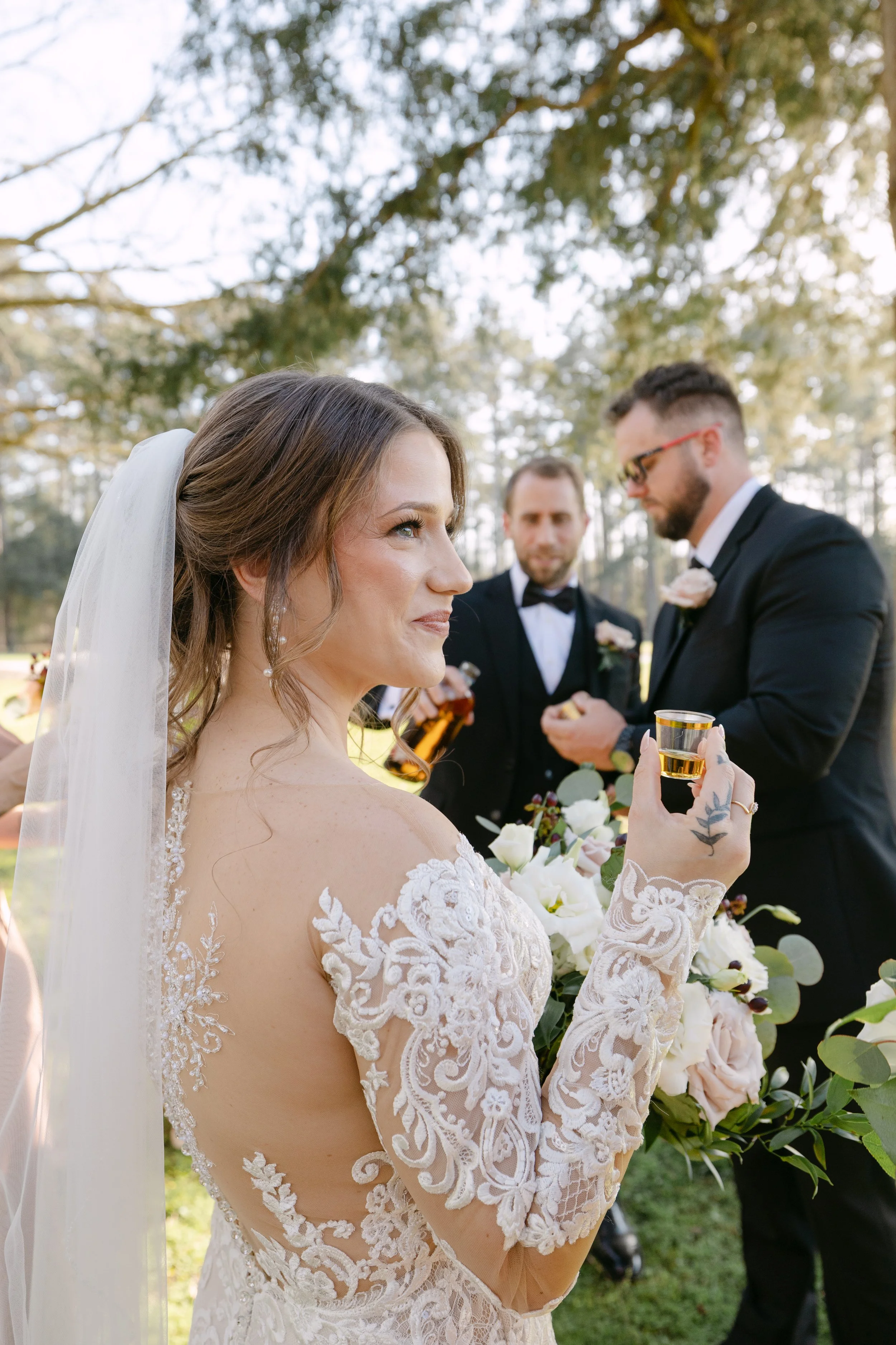 A bride holding a glass of champagne during her outdoor wedding ceremony. She is wearing a lace wedding dress and veil, with a floral bouquet in front of her. Two groomsmen in tuxedos are standing behind her, one holding a bottle, in a wooded natural