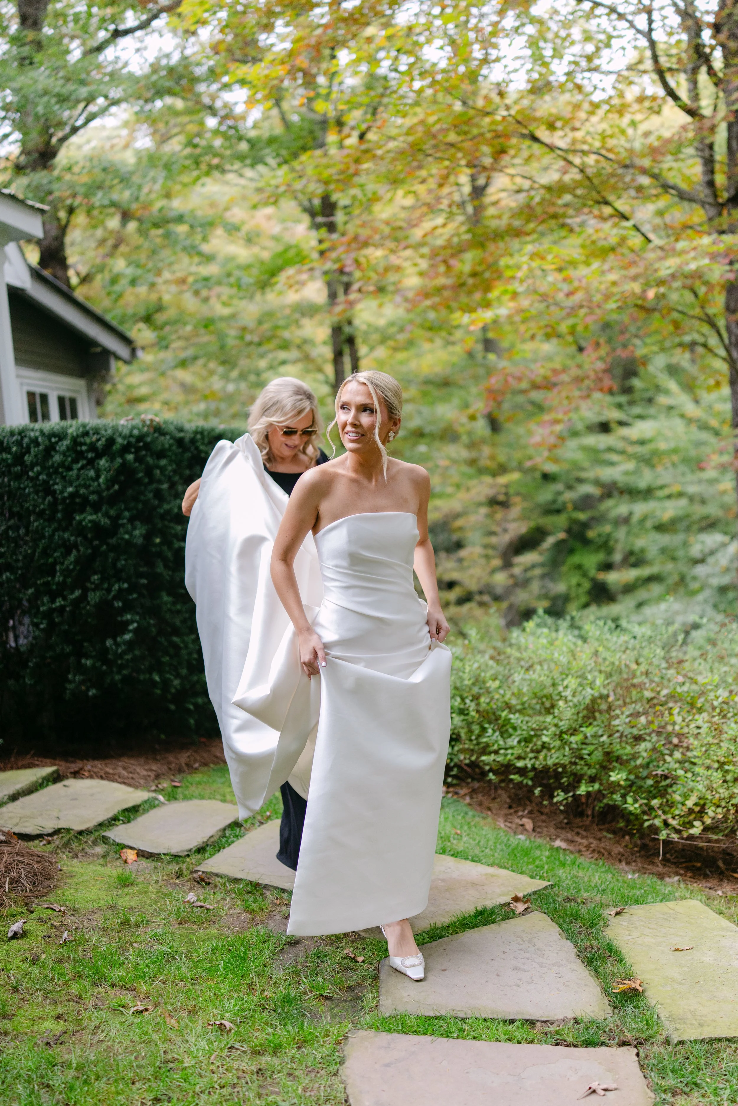 Bride in a strapless white wedding dress walking along a garden path with another woman behind her