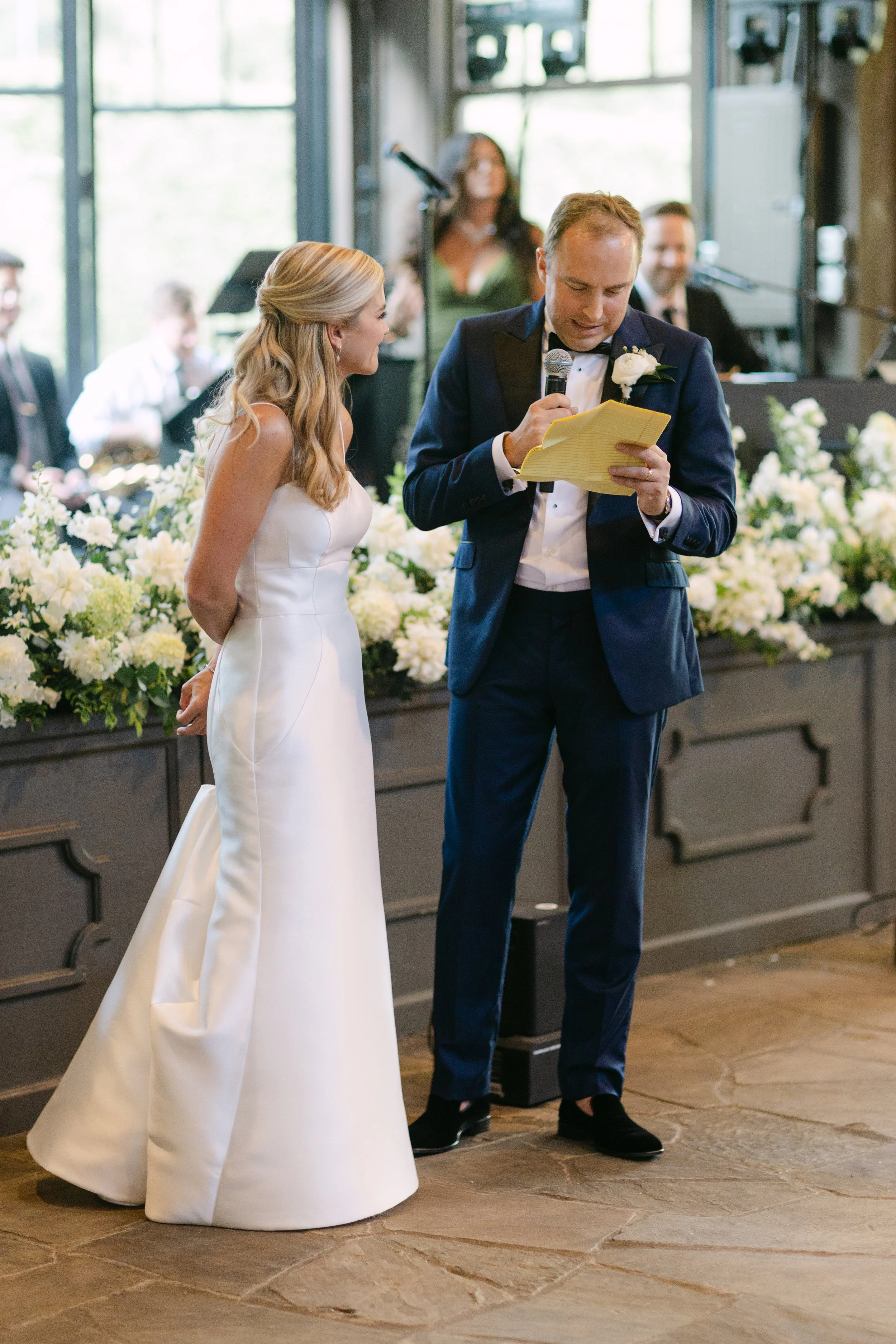 A groom in a navy tuxedo reads vows to a bride in a white wedding gown during a wedding ceremony, with friends and family in the background.