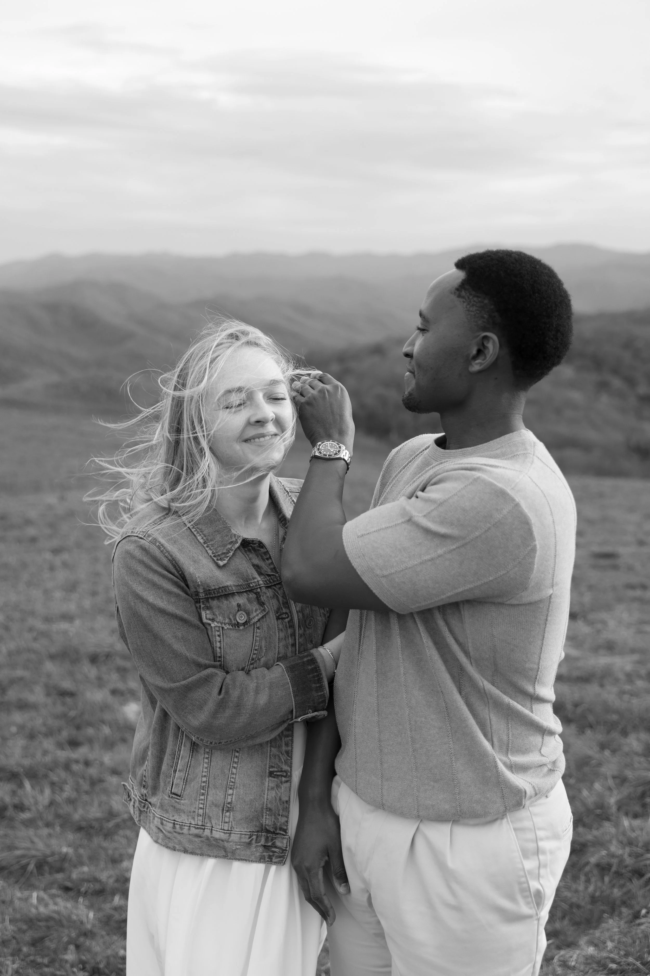 A black and white photo of a woman and a man outdoors, with a landscape of rolling hills and mountains in the background. The woman has long, wavy hair and wears a denim jacket and a long skirt, smiling with her eyes closed. The man has short hair an