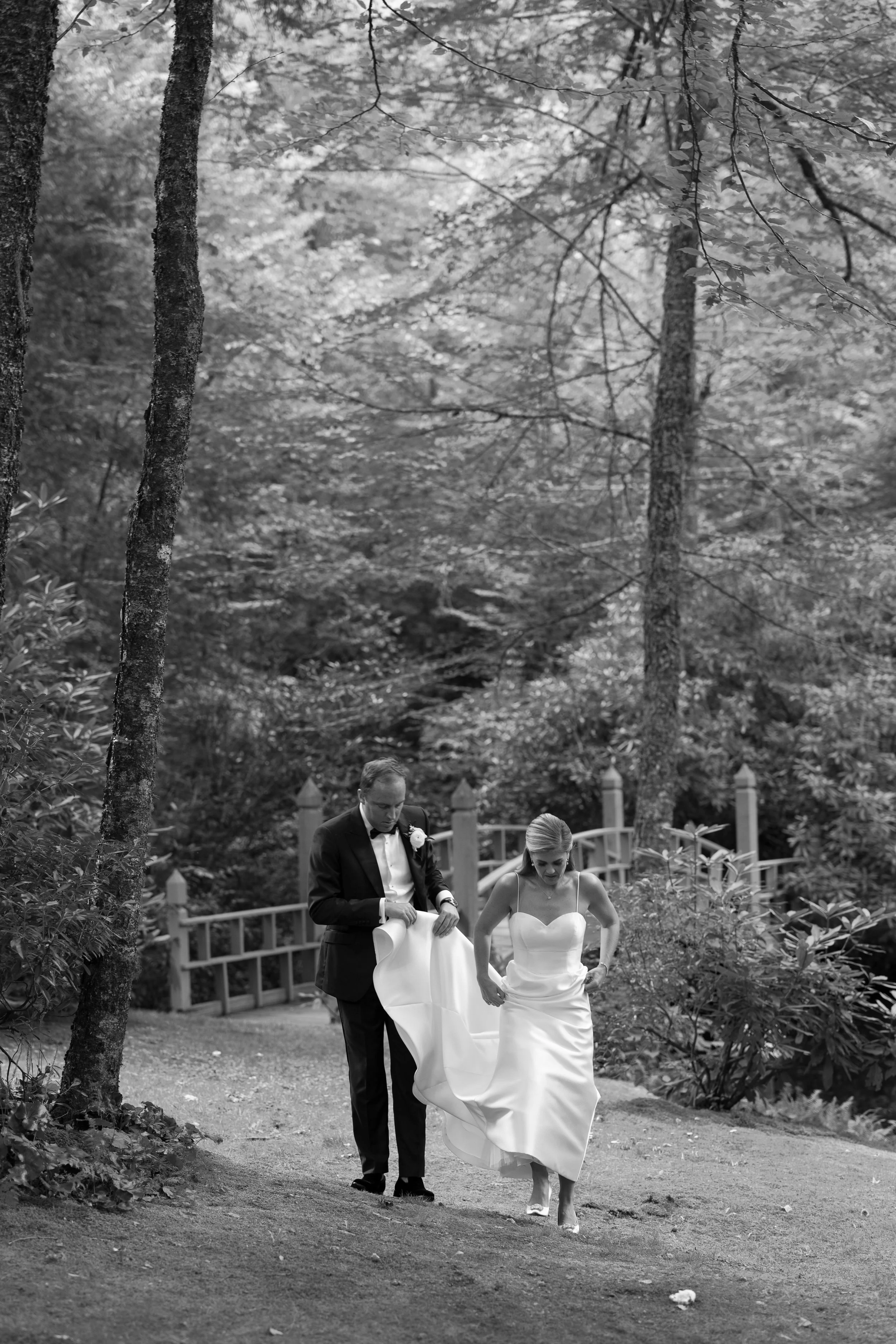 A black and white photo of a bride and groom walking down a garden path. The groom is adjusting his jacket while the bride is holding up her wedding dress. The background shows trees, a wooden fence, and foliage.