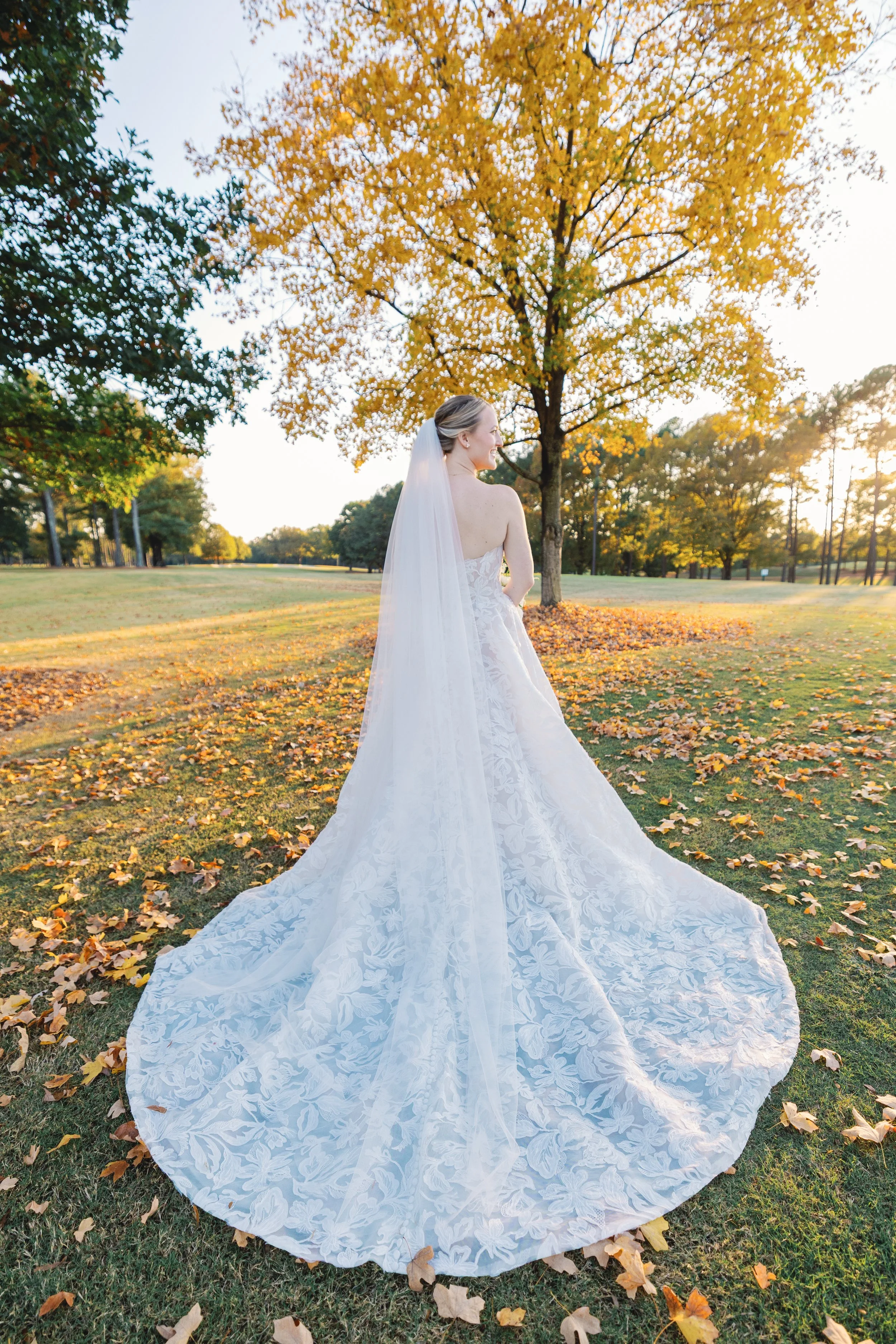 Bride in a white lace wedding gown and veil standing outdoors in a park with yellow autumn trees and fallen leaves.