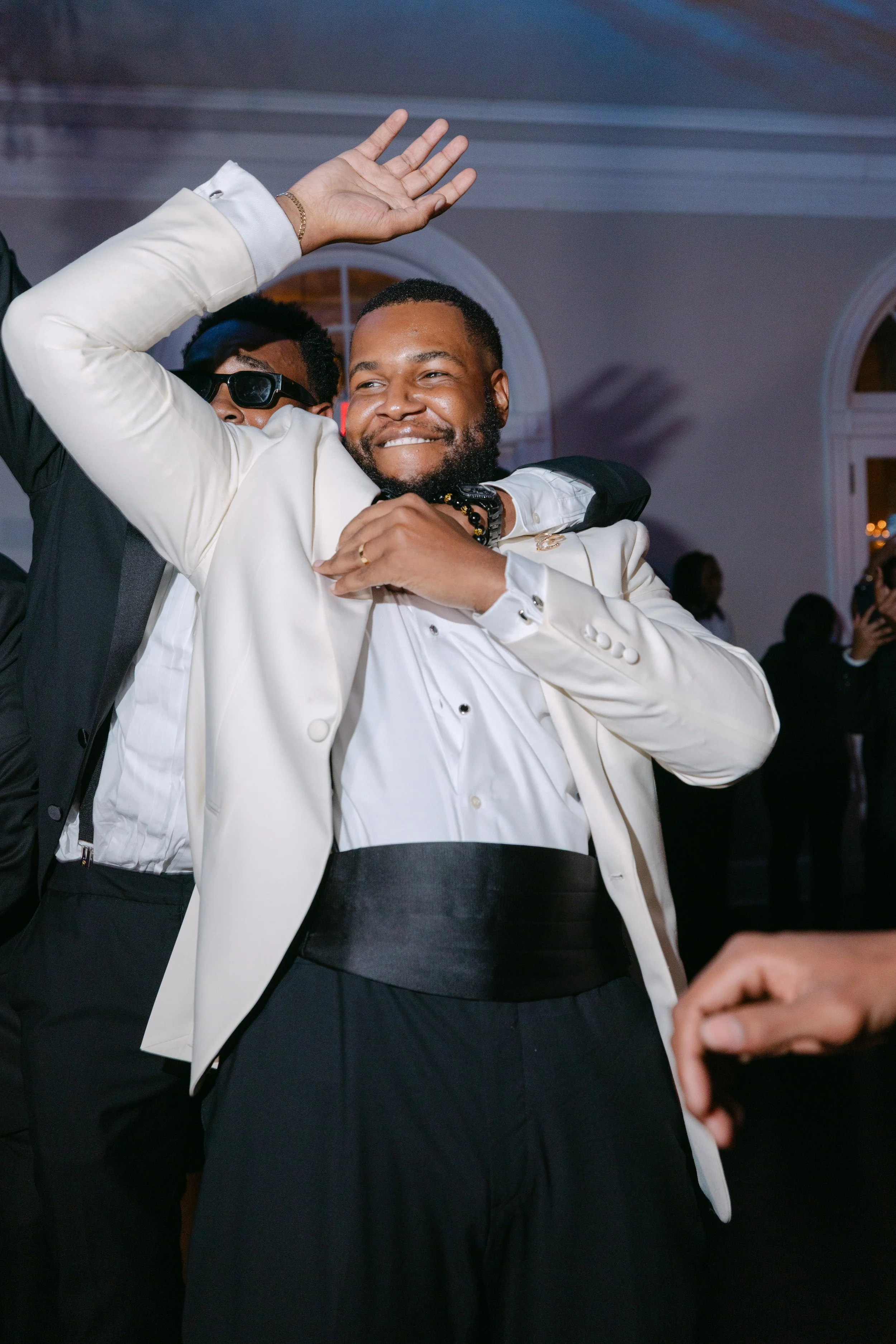 Two men dancing at an indoor event. One man in the foreground is smiling, wearing a cream blazer, black pants, and a beaded bracelet. The other man behind him is wearing dark sunglasses and a black suit.