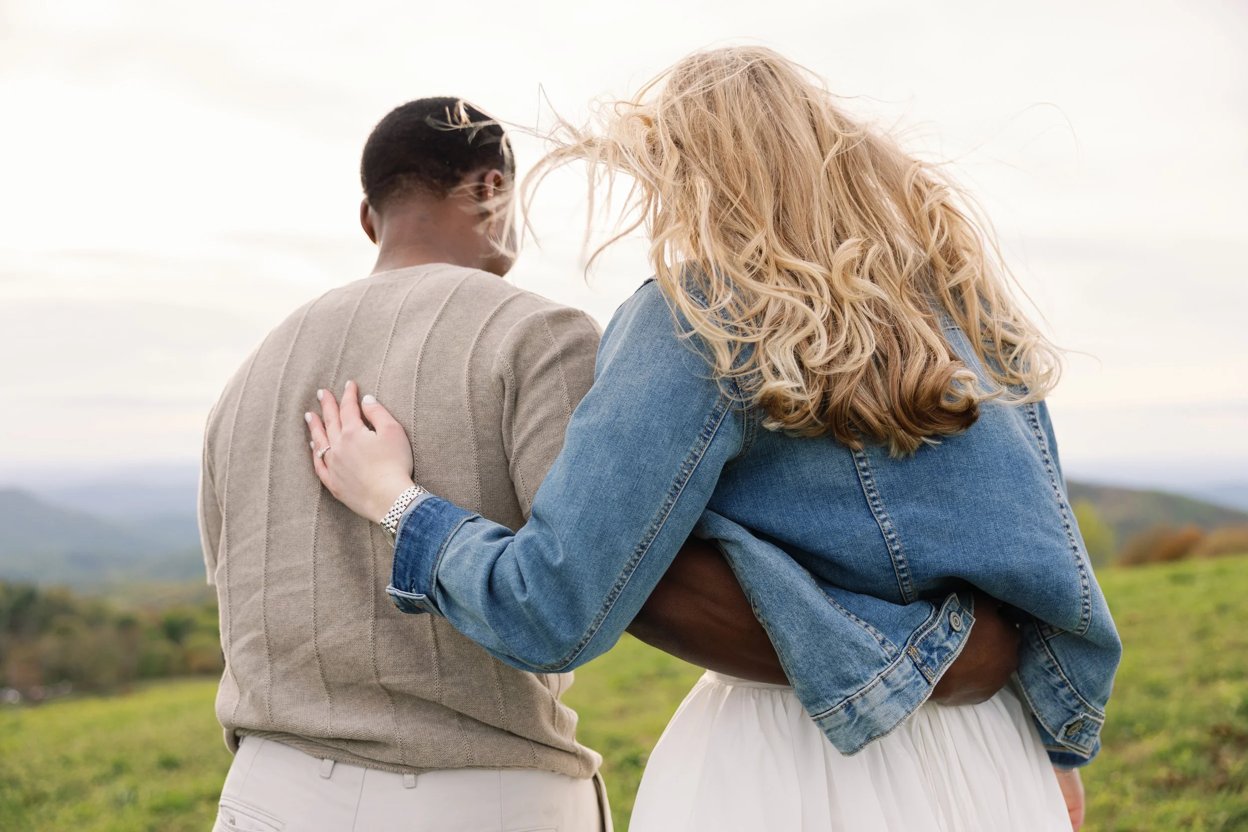 Two people standing outdoors, close together, with one resting their hand on the other's shoulder. The person on the right has long, curly blonde hair and is wearing a denim jacket. The person on the left has short, dark hair and is wearing a beige j