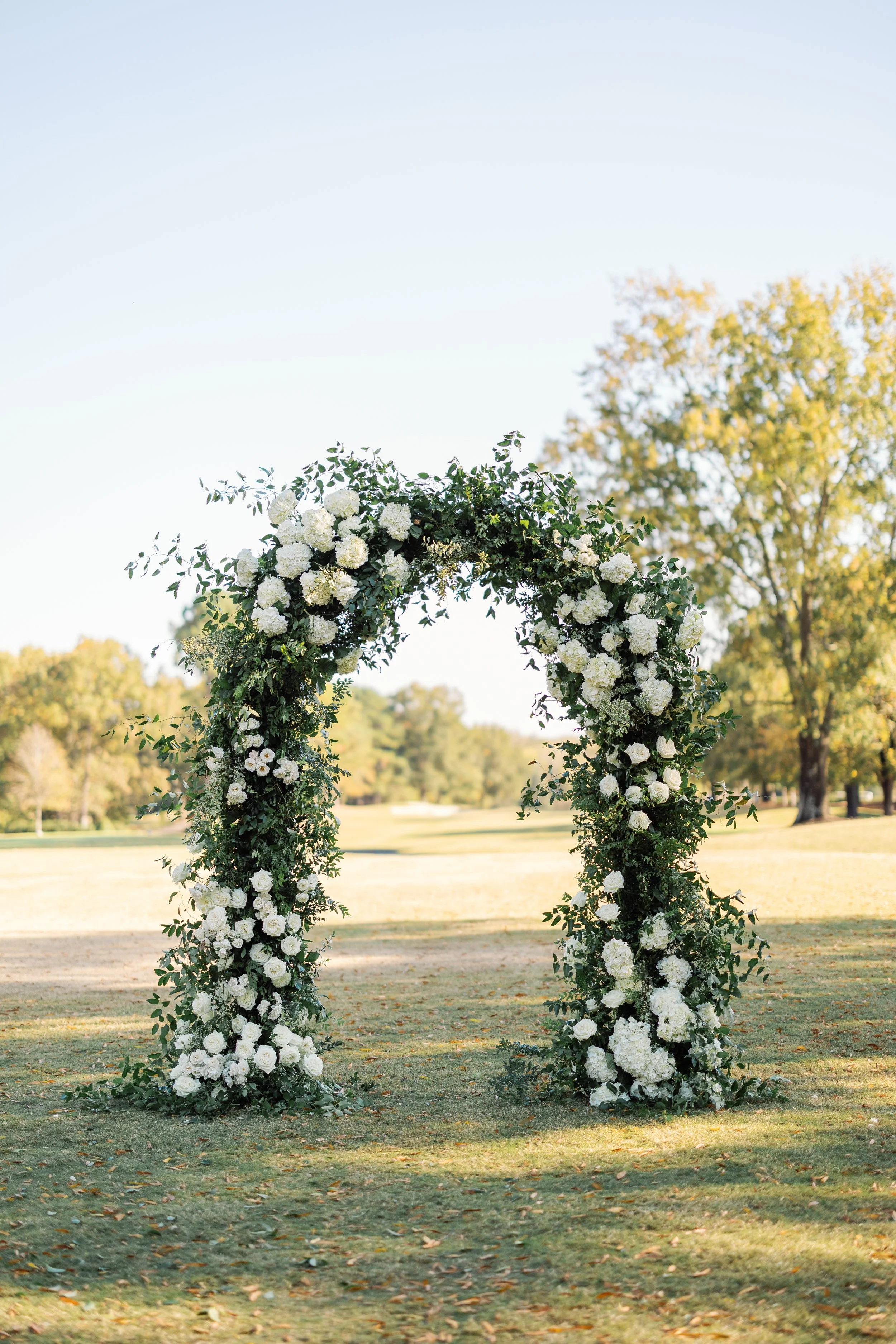 A floral wedding arch decorated with white flowers and greenery, standing outdoors on a grassy field with trees in the background under a clear sky.