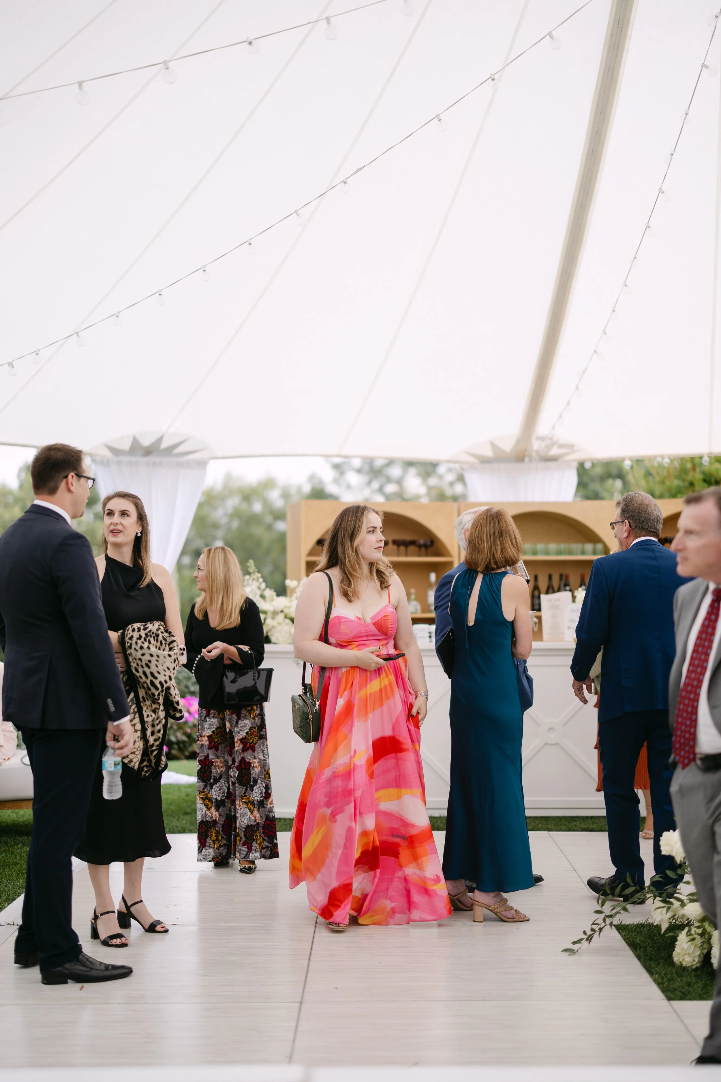 People dressed in formal attire standing and talking under a large white tent at an outdoor event.
