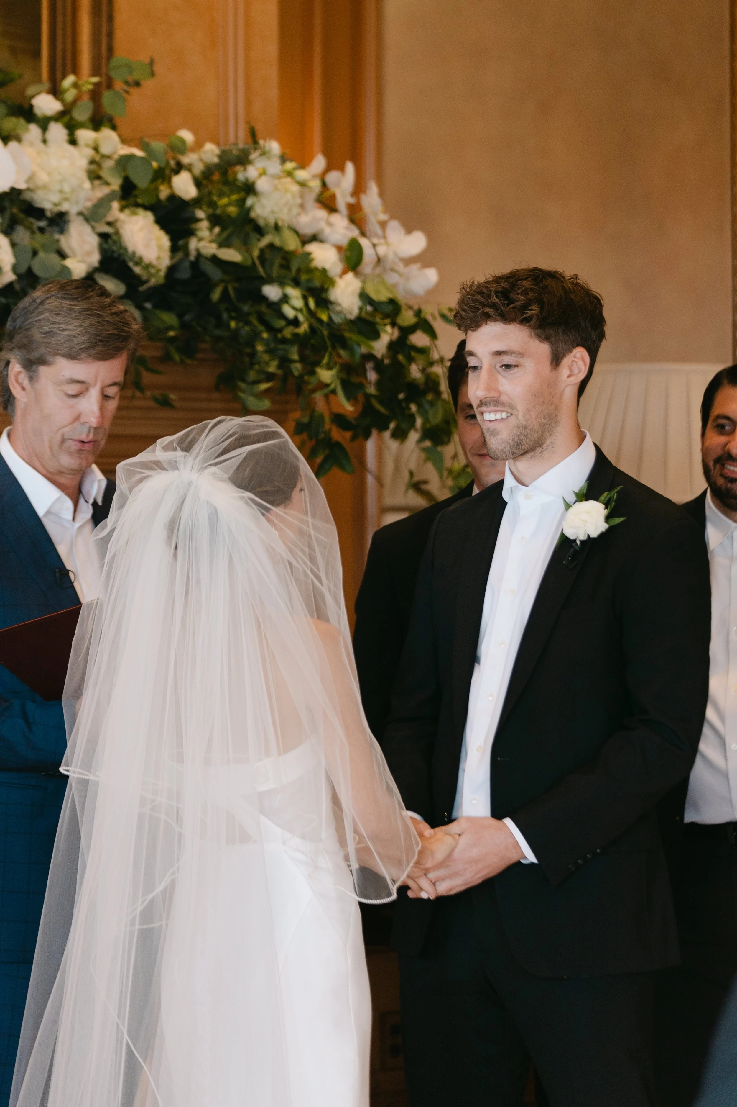 A wedding ceremony with a bride and groom holding hands, exchanging vows, surrounded by officiants and guests, with floral arrangements in the background.