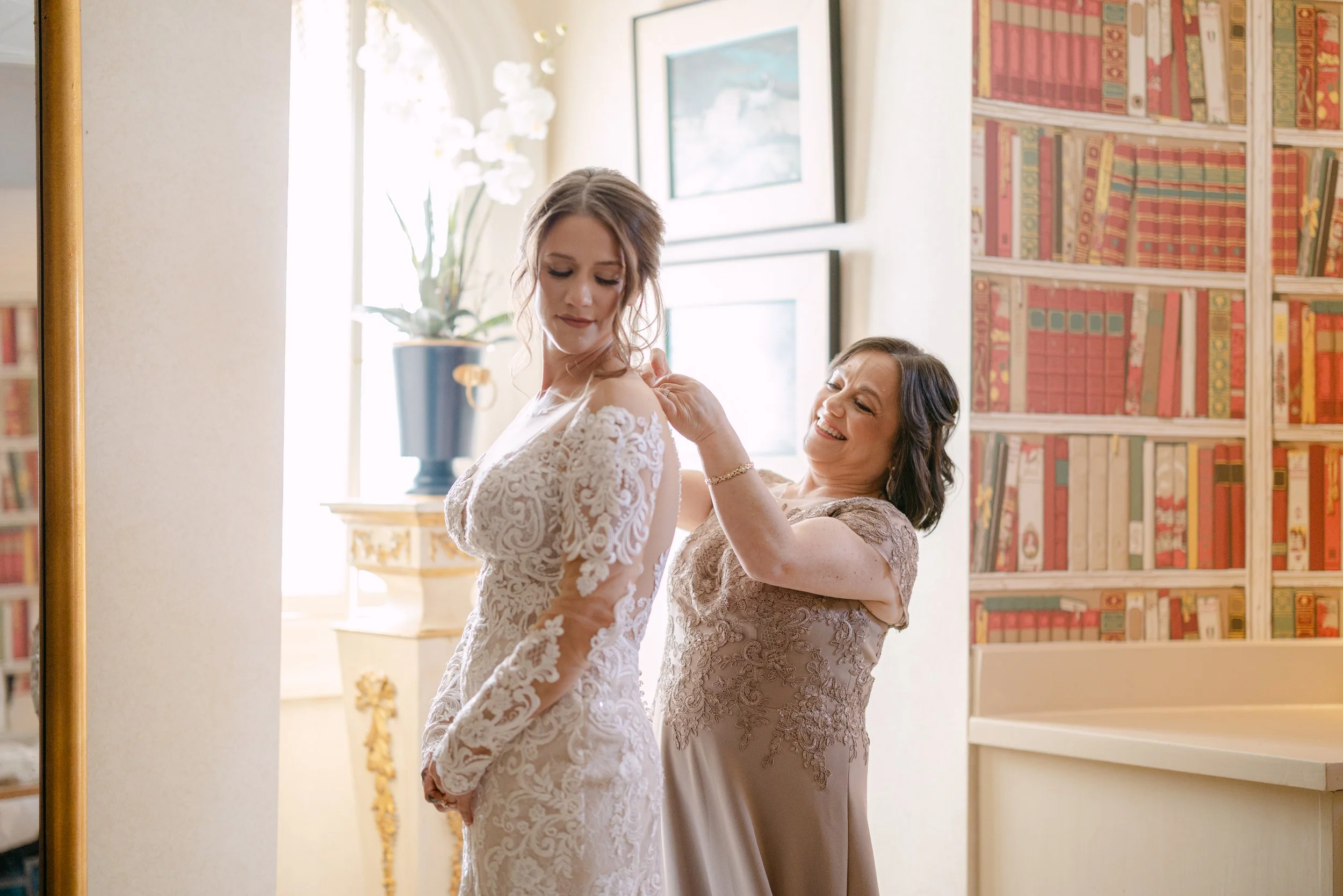 A bride is getting ready, assisted by a woman, in a warmly lit room decorated with framed pictures and a bookshelf.