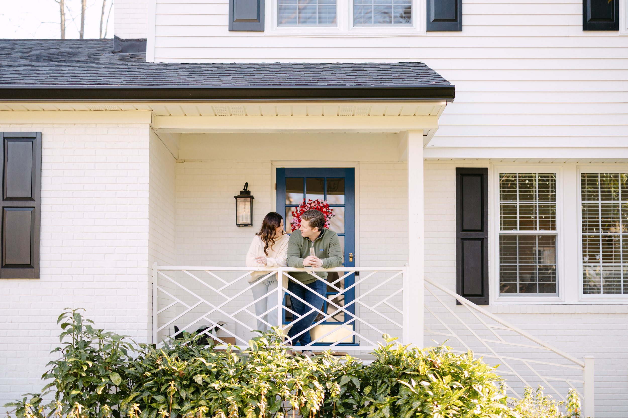 A couple stands on the front porch of a white house with black window shutters, sharing a moment under a holiday wreath on the door.
