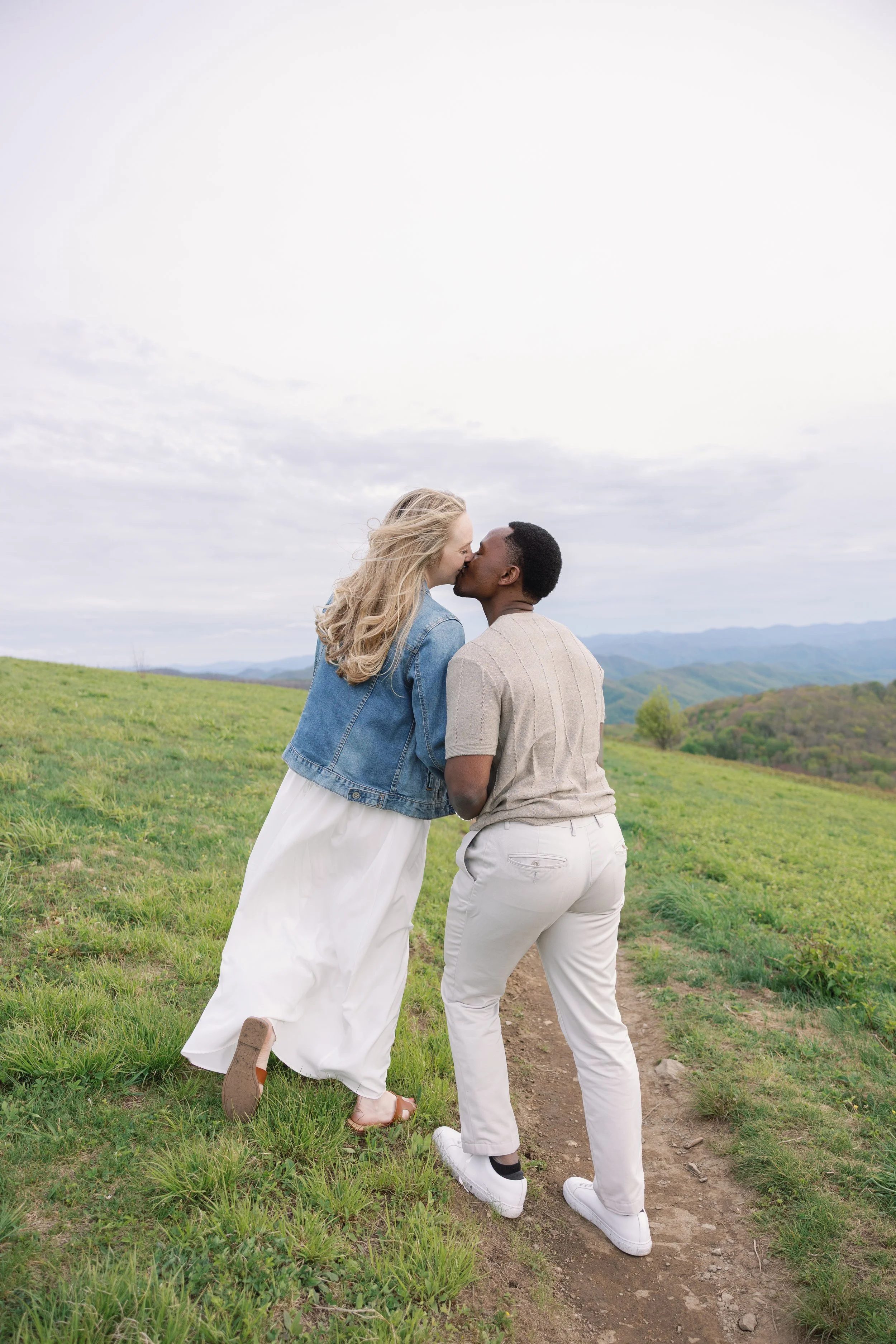 A couple kissing outdoors in a green field with rolling hills in the distance, cloudy sky overhead.