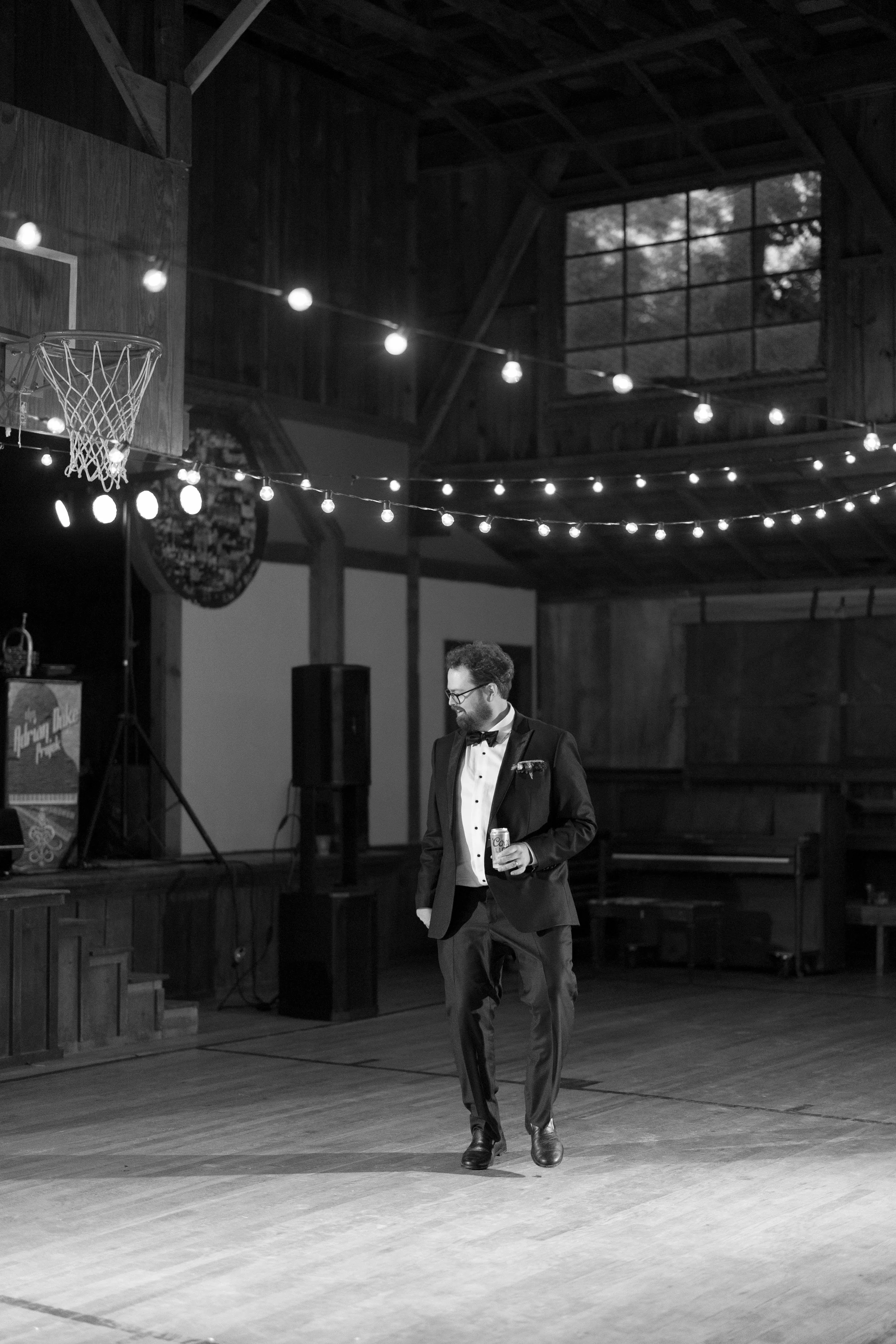 A man in a tuxedo holding a drink, standing alone in a dimly lit wooden indoor venue decorated with string lights, with a basketball hoop in the background.