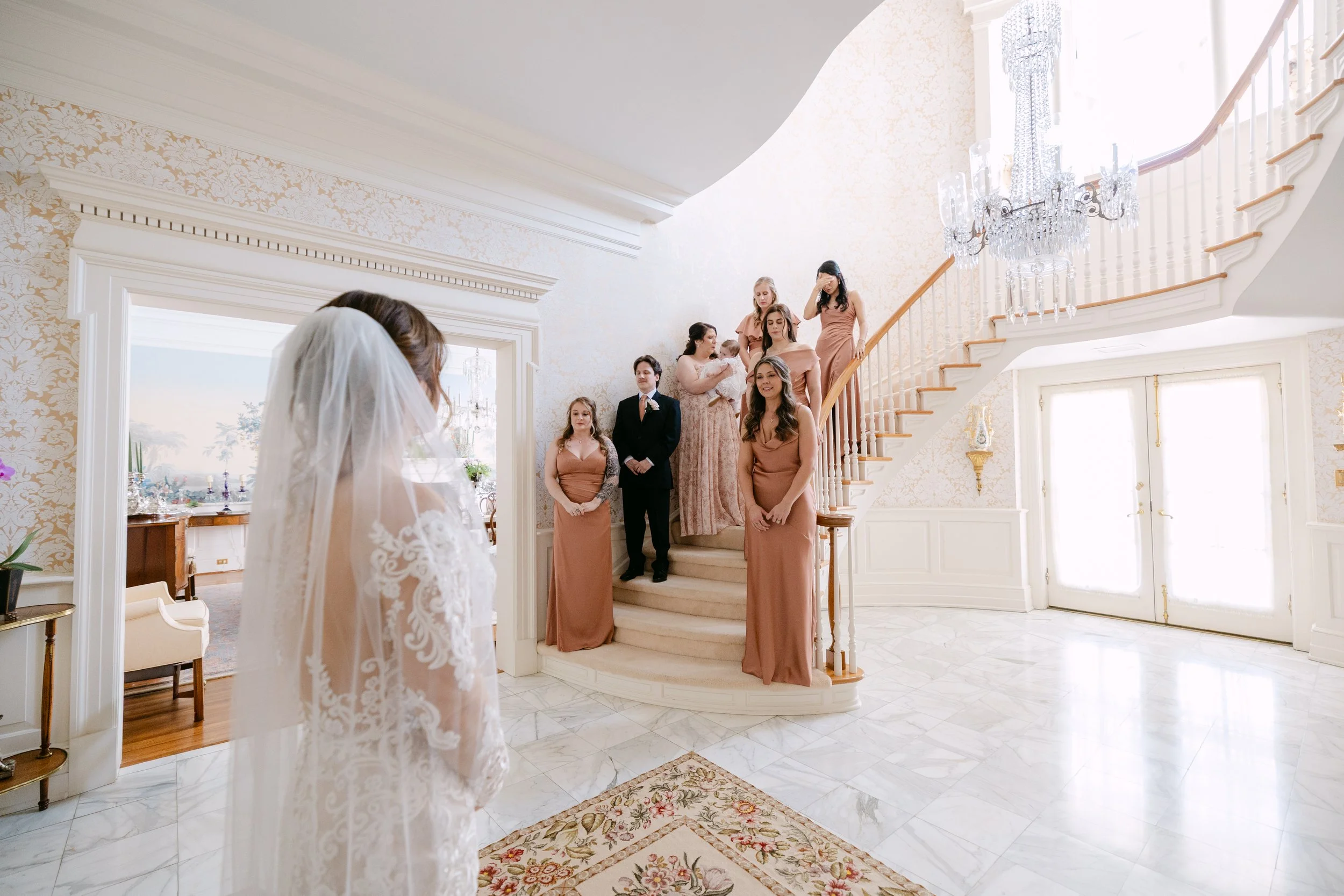 A bride with a veil facing a group of bridesmaids and a man in formal attire in a bright, elegant room with a marble floor, ornate wallpaper, and a grand staircase.