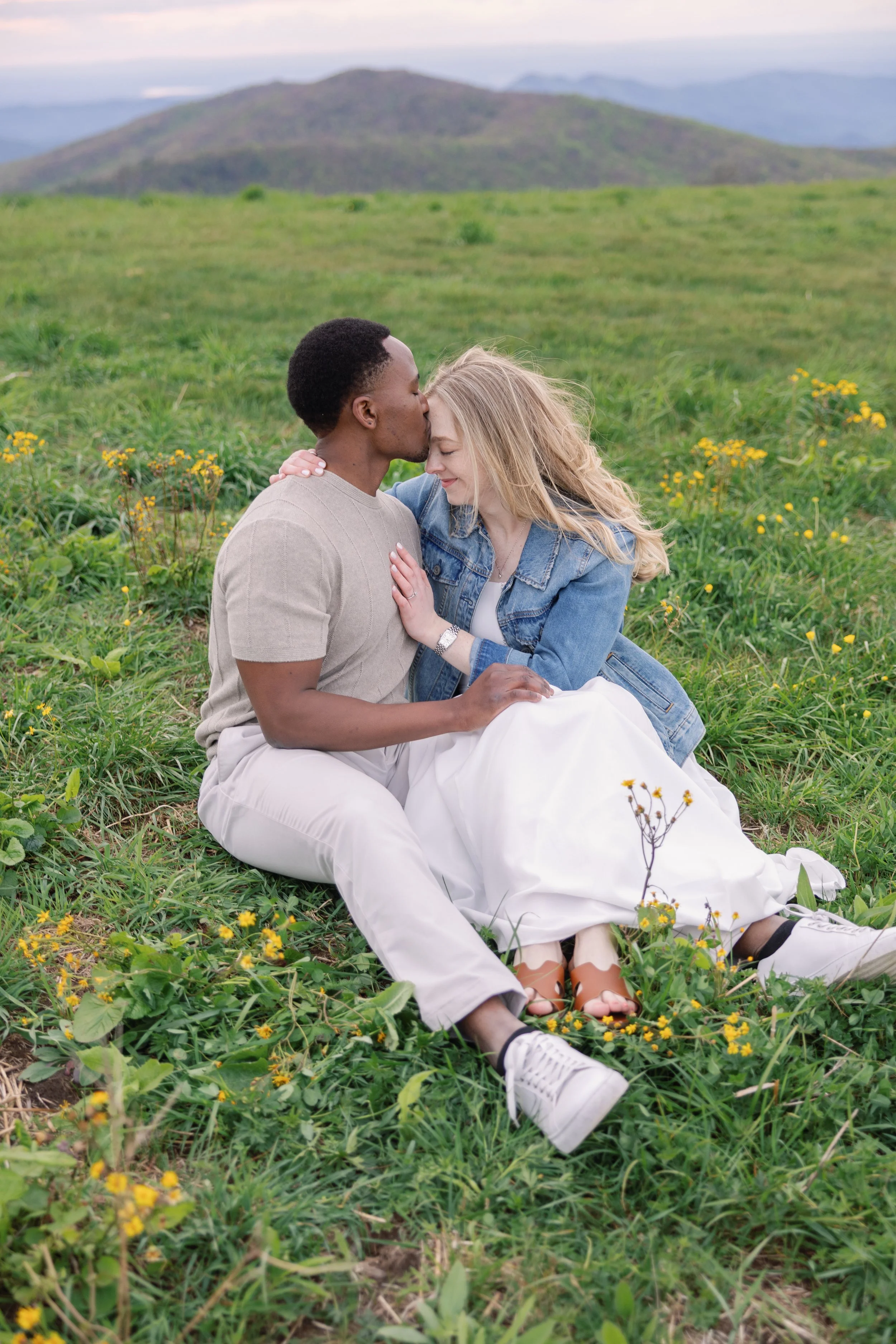 A couple sits in a grassy field with yellow wildflowers; the man, with short dark hair, is kissing the woman's forehead, and she has long blonde hair. The background features distant mountains under a pastel sky.