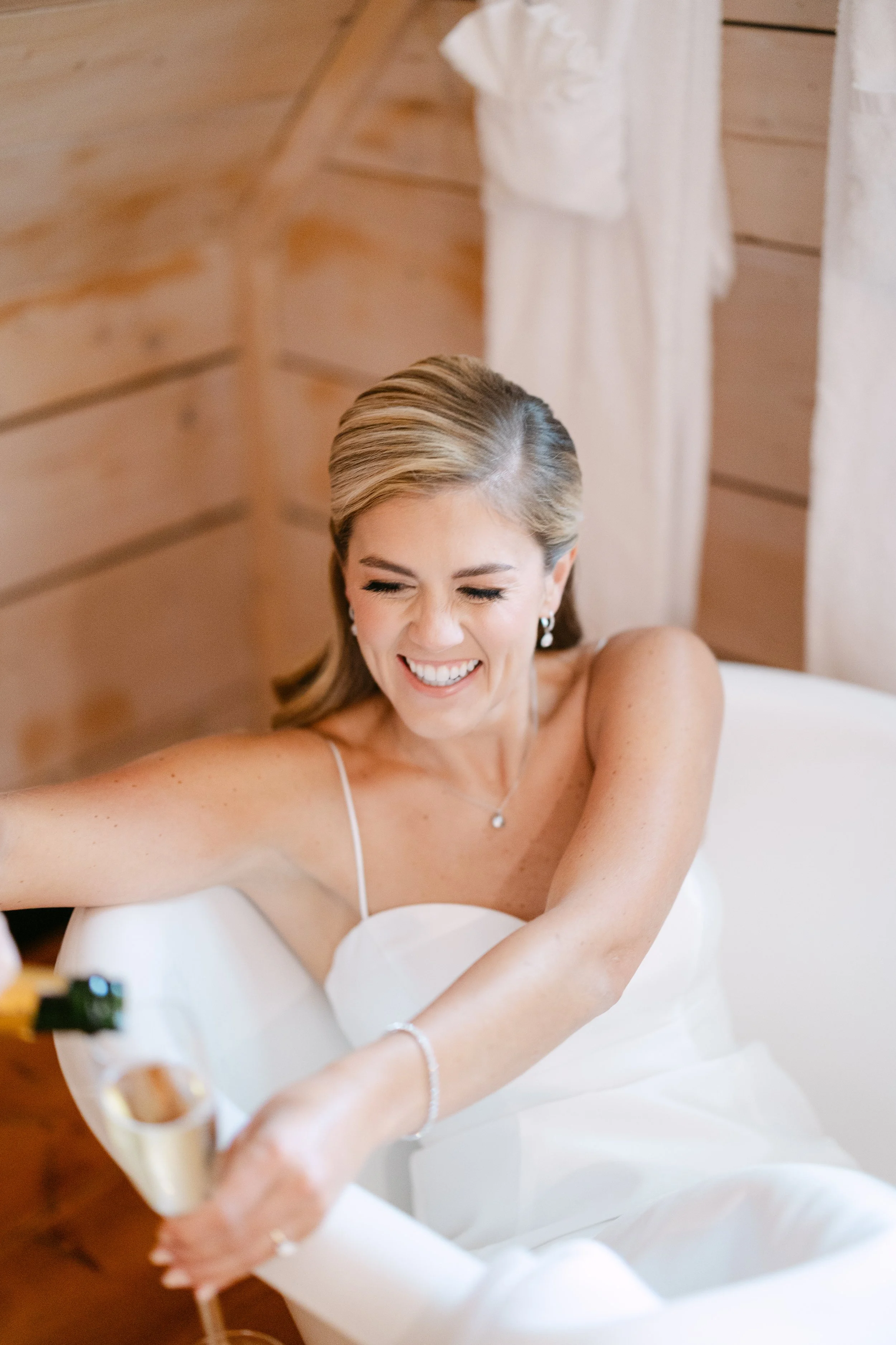 A bride laughing and smiling while sitting in a white bathtub, holding a glass of champagne.