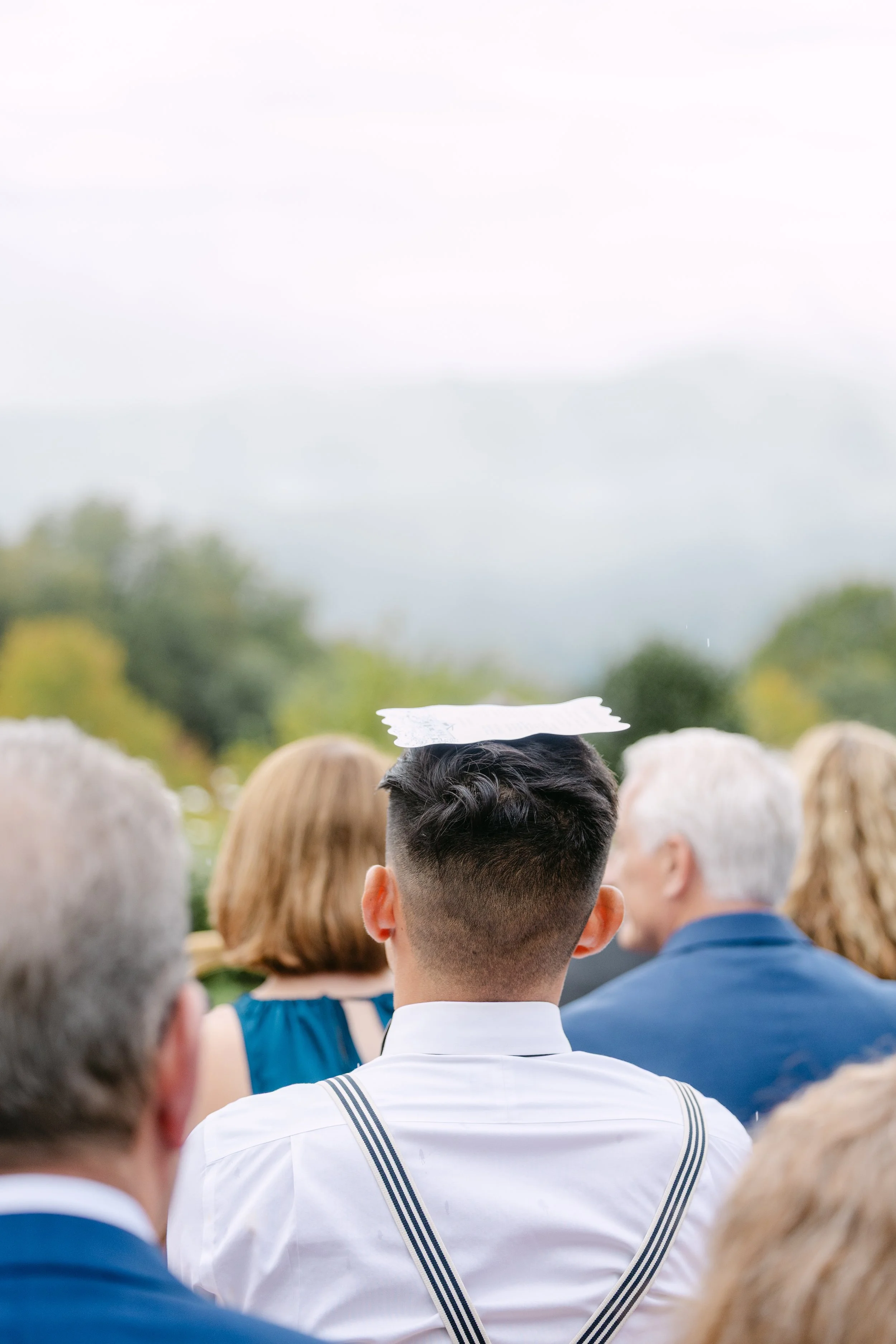 A young man in a white dress shirt with suspenders, seen from behind, attending an outdoor graduation ceremony during daytime, with a mortarboard in his hand and trees in the background.