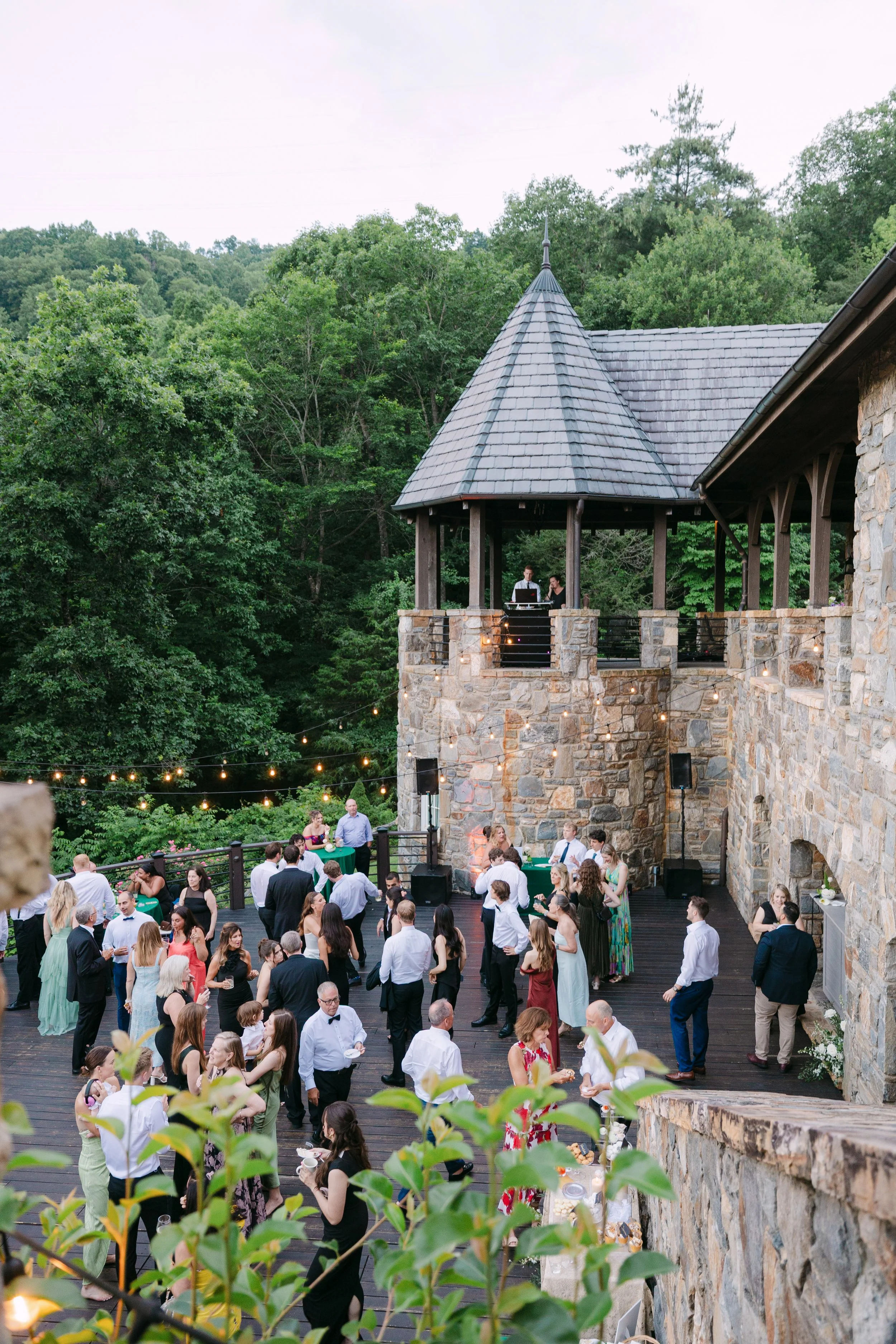 Guests dancing and socializing on a wooden outdoor terrace at a gathering or celebration, surrounded by trees and string lights, with a stone and wood building in the background.